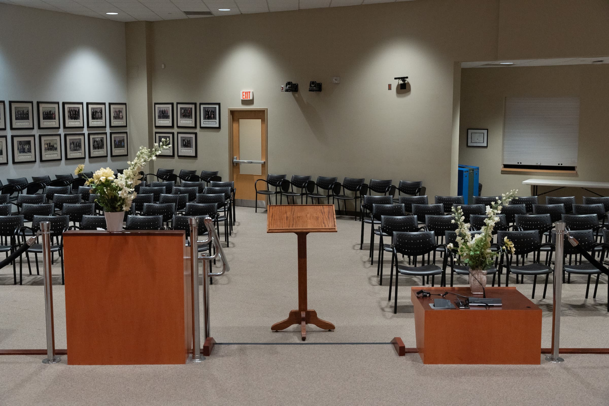 A small conference room with rows of empty chairs, a lectern, and floral arrangements on wooden stands.