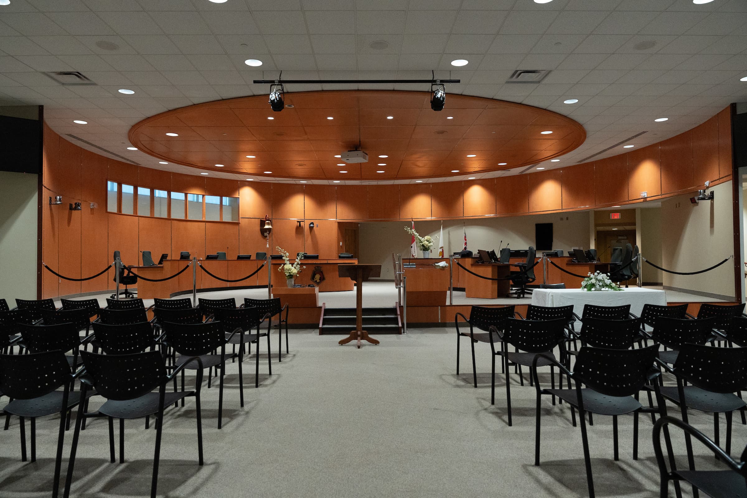 A large, empty courtroom with rows of black chairs facing a wooden judge's bench and several monitors.