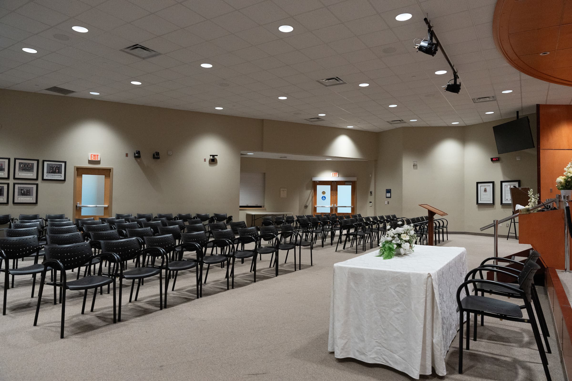 Rows of empty chairs face a table with a white cloth and flowers in a large, well-lit conference room.