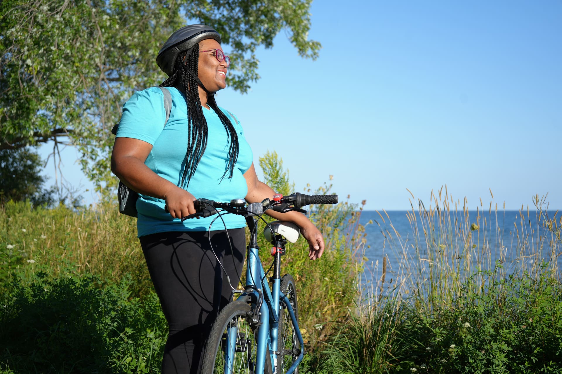Person wearing a helmet stands next to a bicycle by a lakeside, surrounded by greenery on a sunny day.