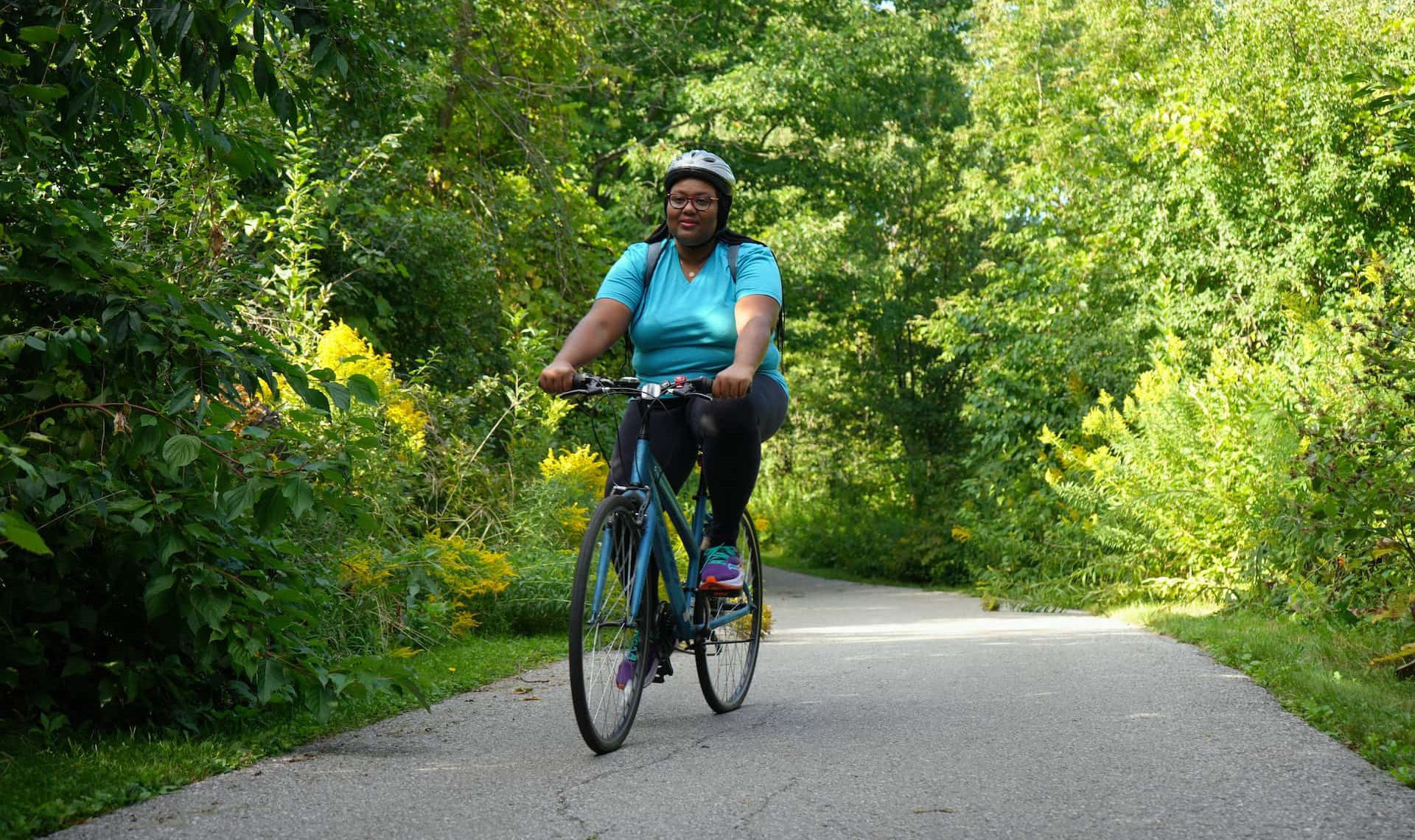 A person wearing a helmet rides a bicycle on a paved path surrounded by green trees and bushes.
