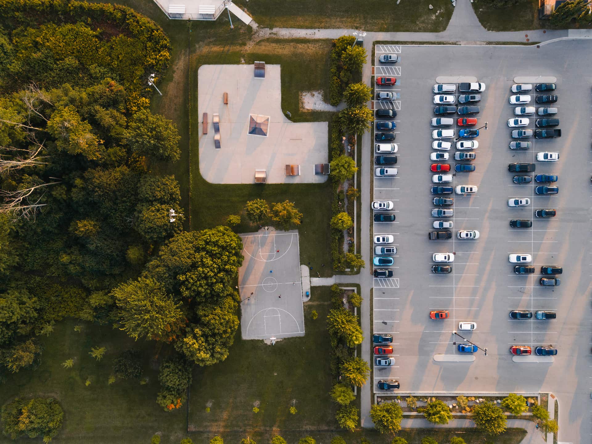 Aerial view of a car park, basketball court, and playground surrounded by grass and trees.