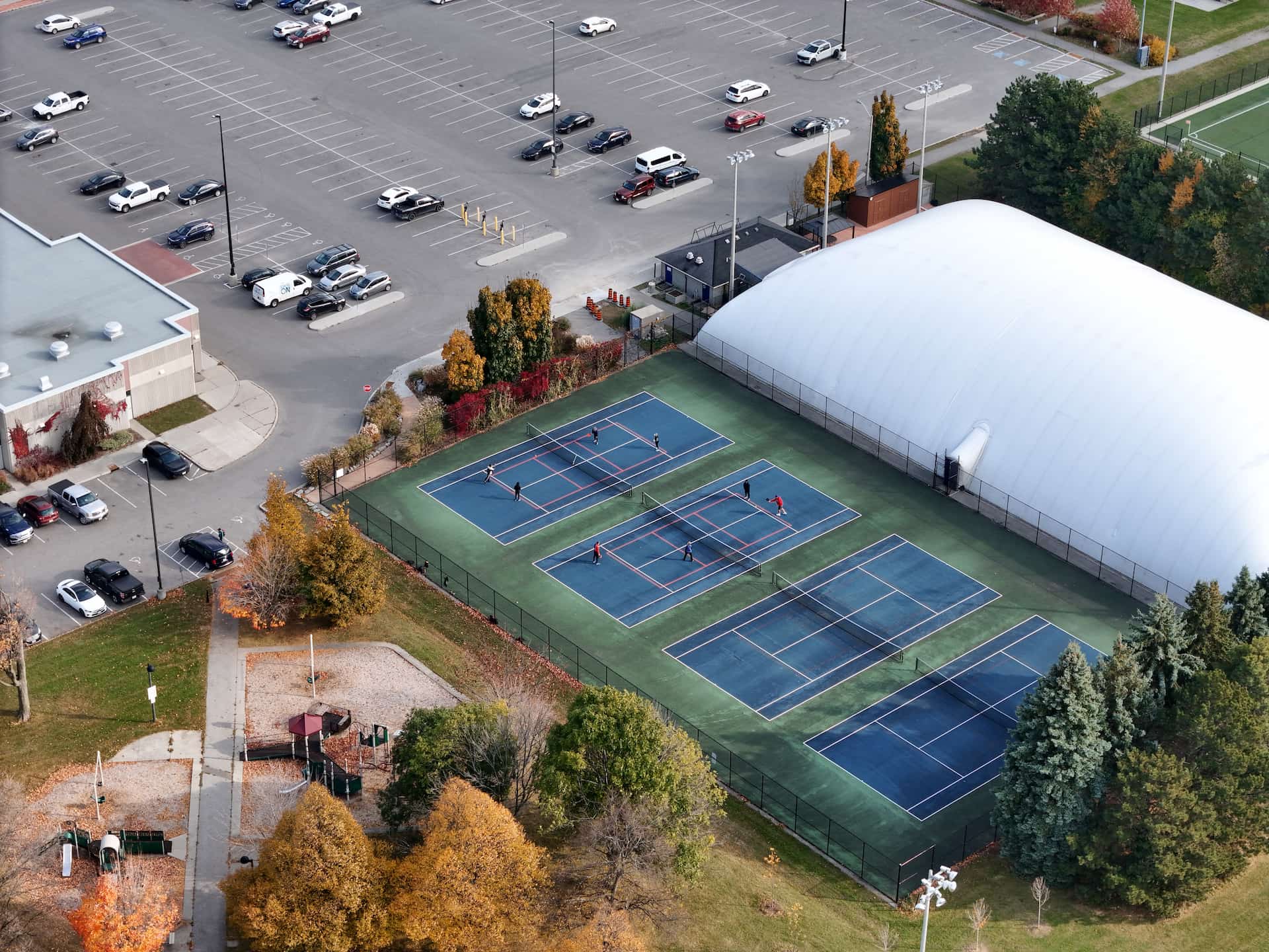 Aerial view of outdoor tennis courts with players, adjacent car park, playground, and an indoor sports centre.