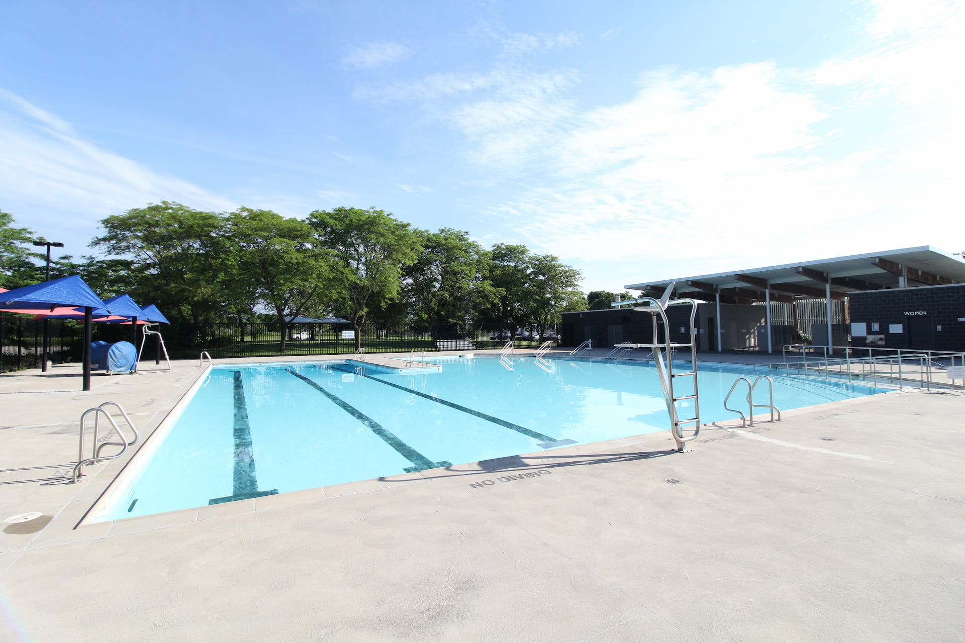 Outdoor swimming pool with clear water, empty decking, ladders, and shaded seating area under a sunny sky.