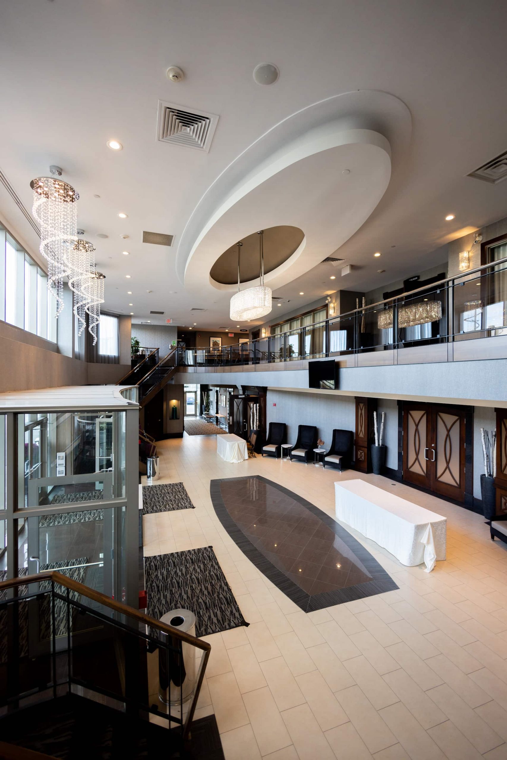 Spacious modern hotel lobby with chandeliers, glass lift, white tiled floor, and reception desks along the walls.