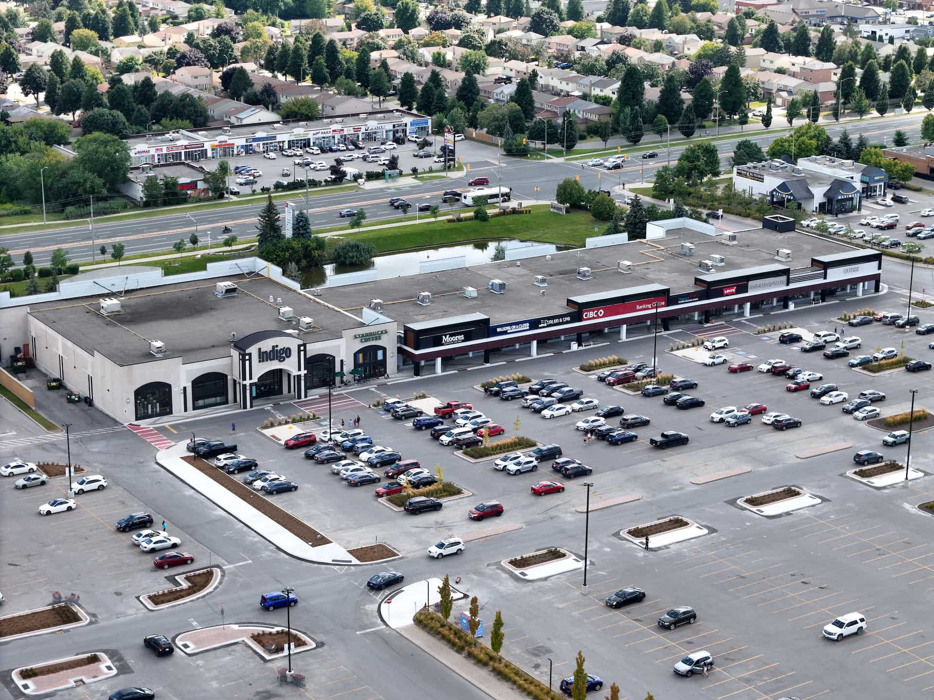 Aerial view of a shopping precinct with several shops and a car park containing many cars, with roads and houses nearby.