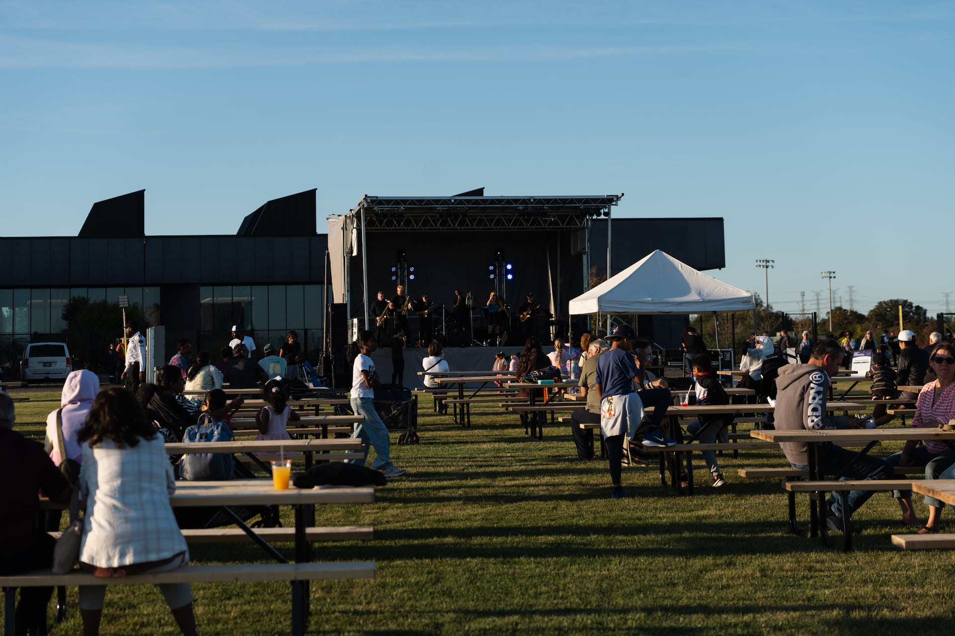 People sit at picnic tables on a lawn, facing an outdoor stage with a band performing in front of a modern building.