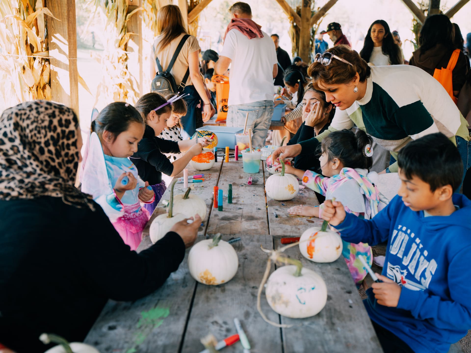 Children and adults sit at a wooden table painting white pumpkins during an outdoor autumn craft activity.