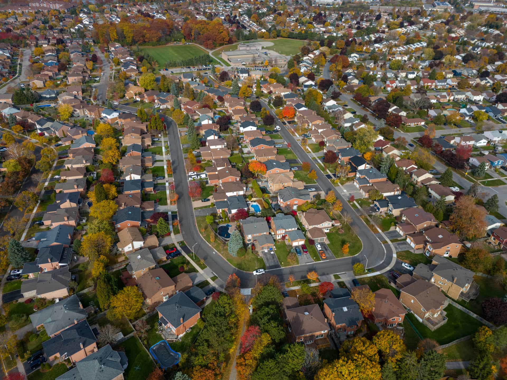 Aerial view of a suburban neighbourhood with houses, streets, trees, and a school surrounded by autumn foliage.