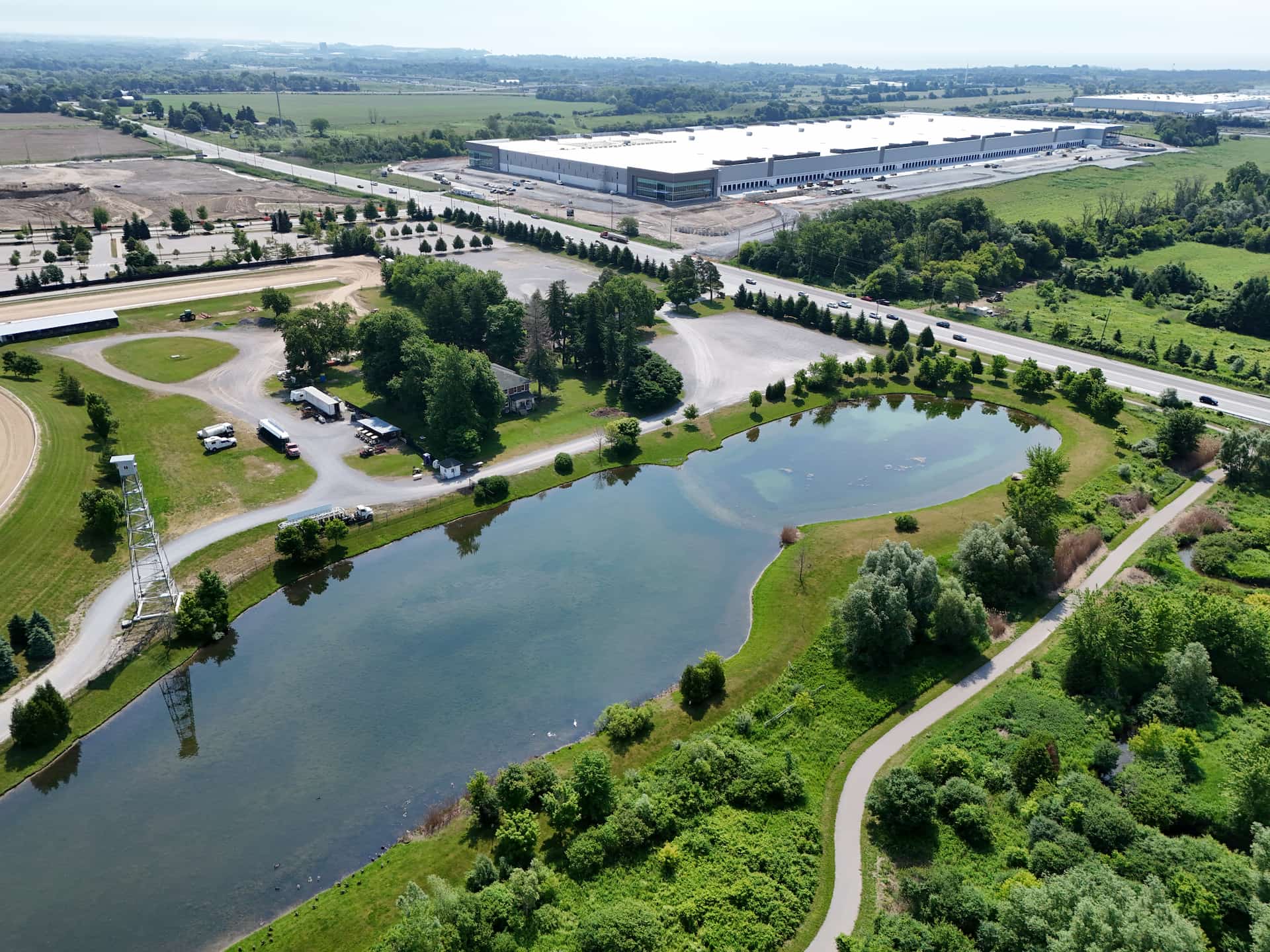Aerial view of a park with a pond, trees, car park, and a large industrial building in the background.