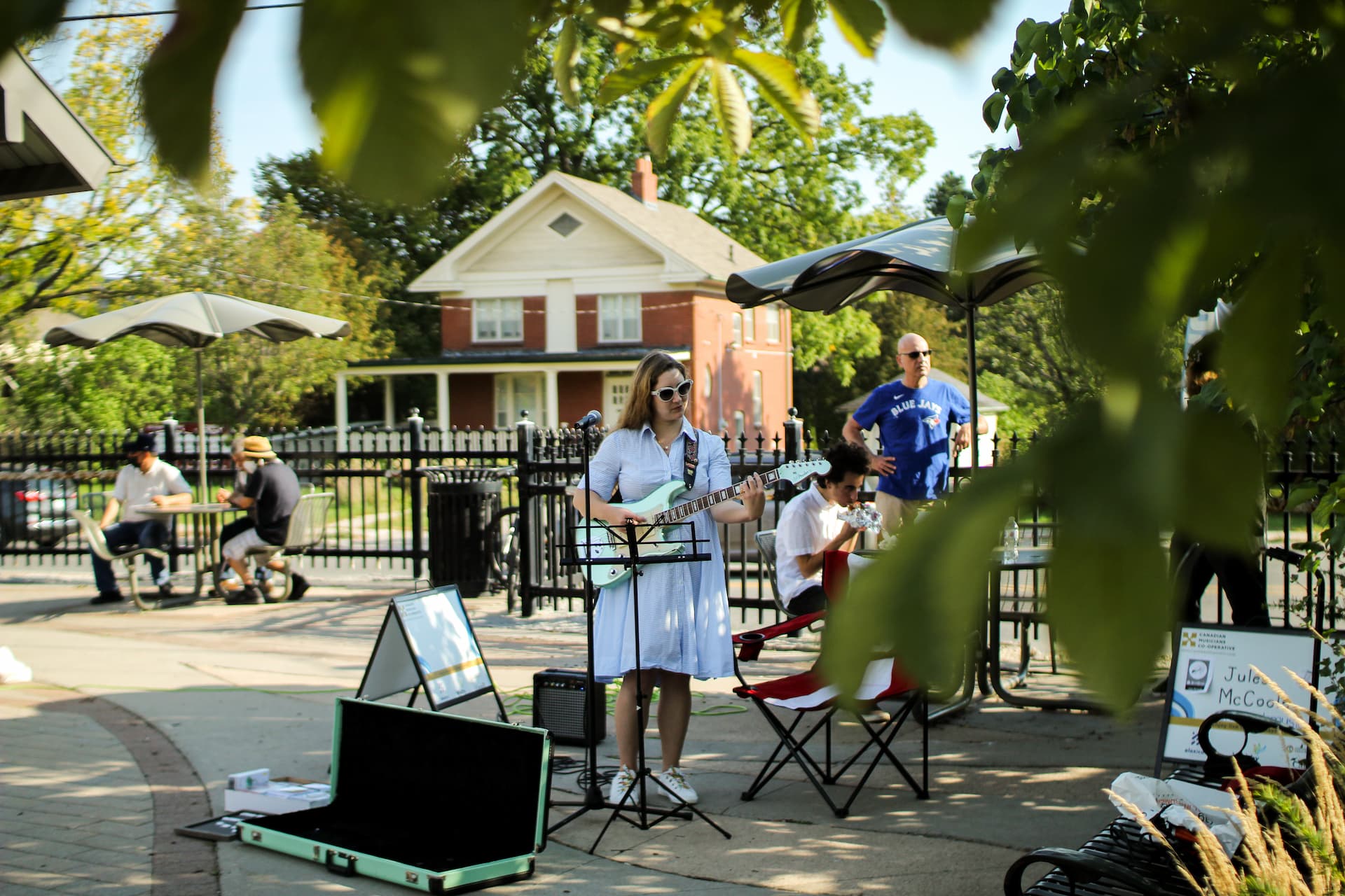 A woman plays electric guitar and sings outdoors near a house, with people sitting at tables and listening.
