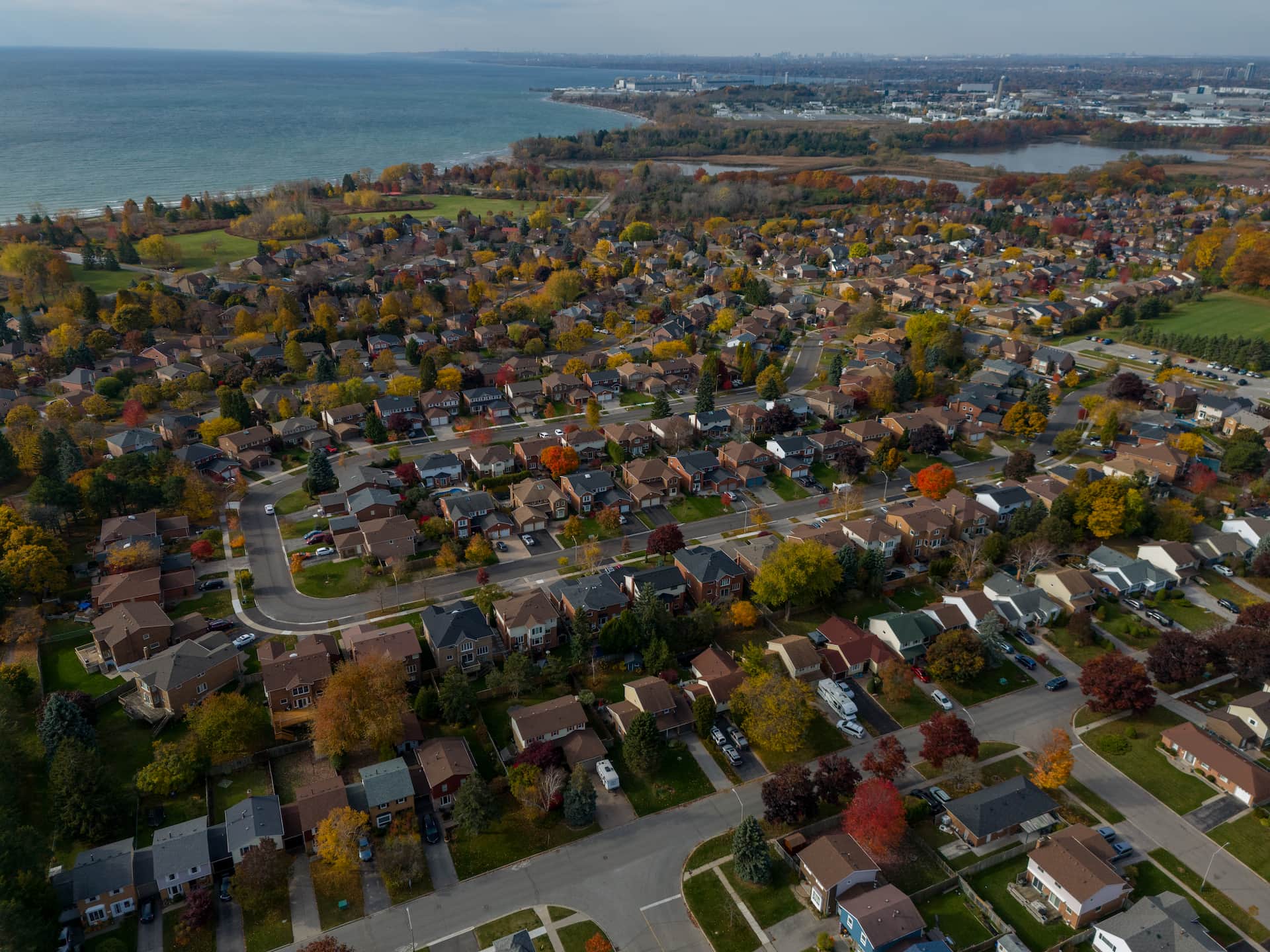 Aerial view of a suburban neighbourhood with rows of houses, trees with autumn foliage, and a body of water in the distance.