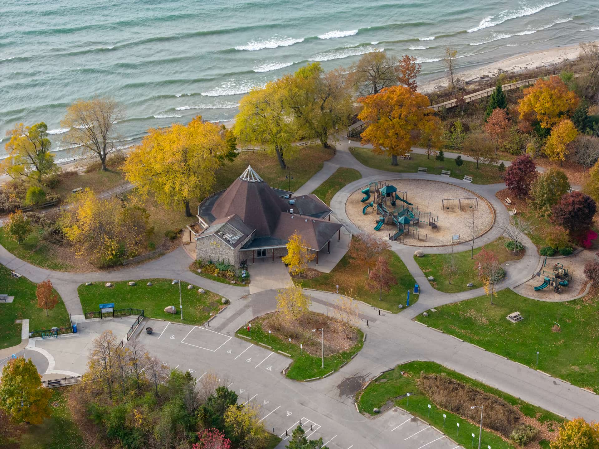 Aerial view of a park with a building, playgrounds, walking paths, autumn trees, and a nearby shoreline.
