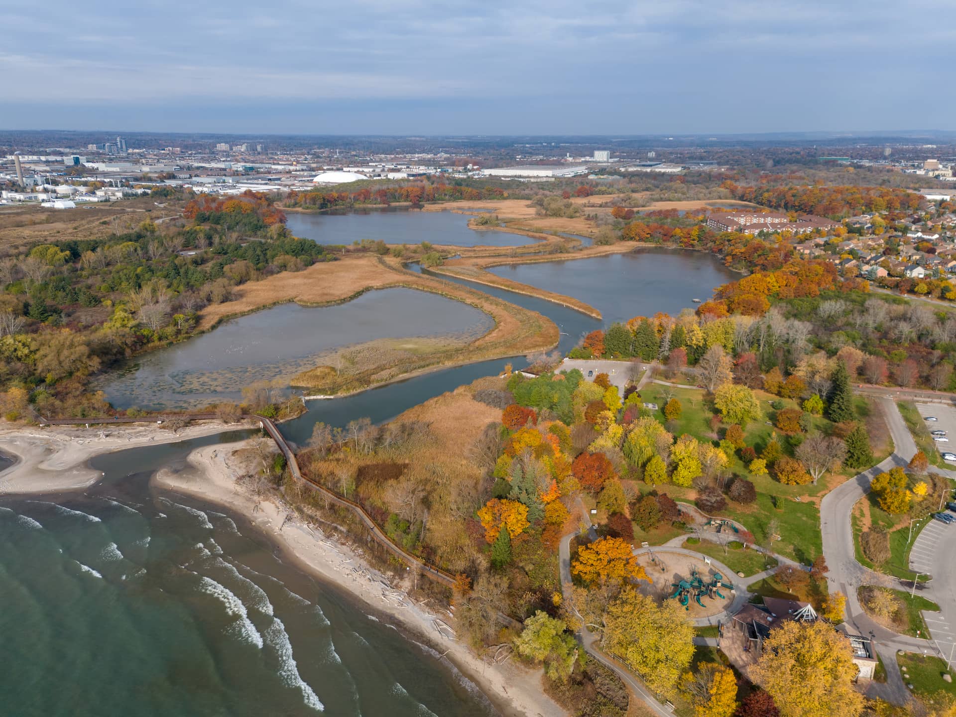 Aerial view of a coastal park with ponds, trees in autumn colours, and a nearby urban area under a cloudy sky.
