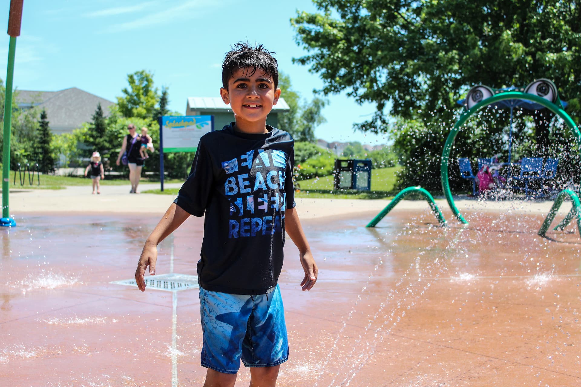 A boy in a black shirt and blue shorts stands smiling at a splash park, with water spraying around him on a sunny day.