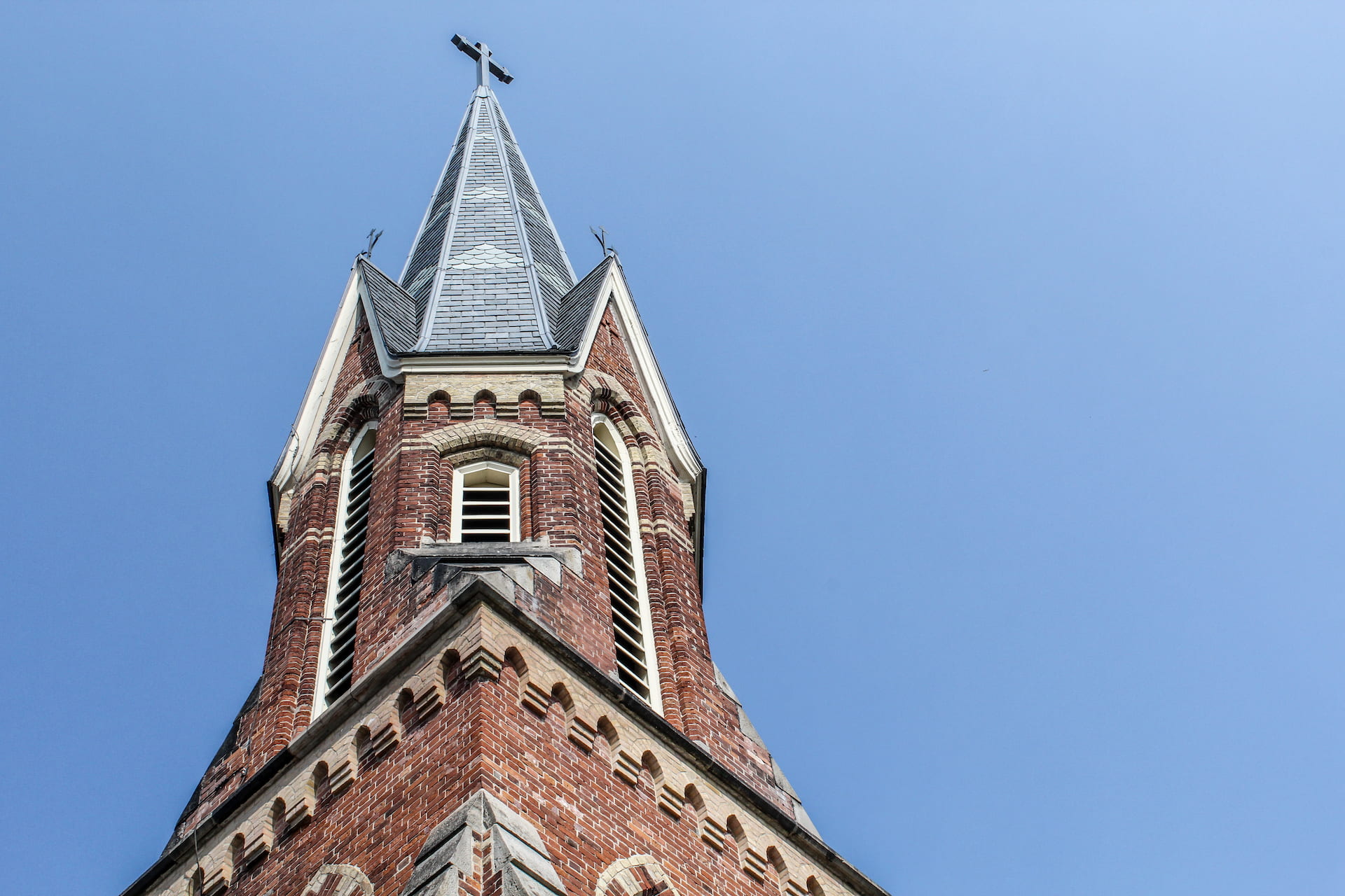 Brick church spire with a cross on top against a clear blue sky.