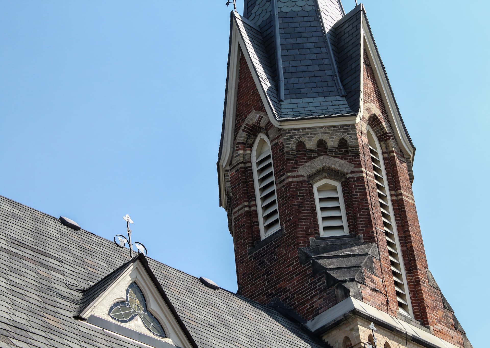 Close-up of a brick church spire with arched windows and a slate roof against a clear blue sky.
