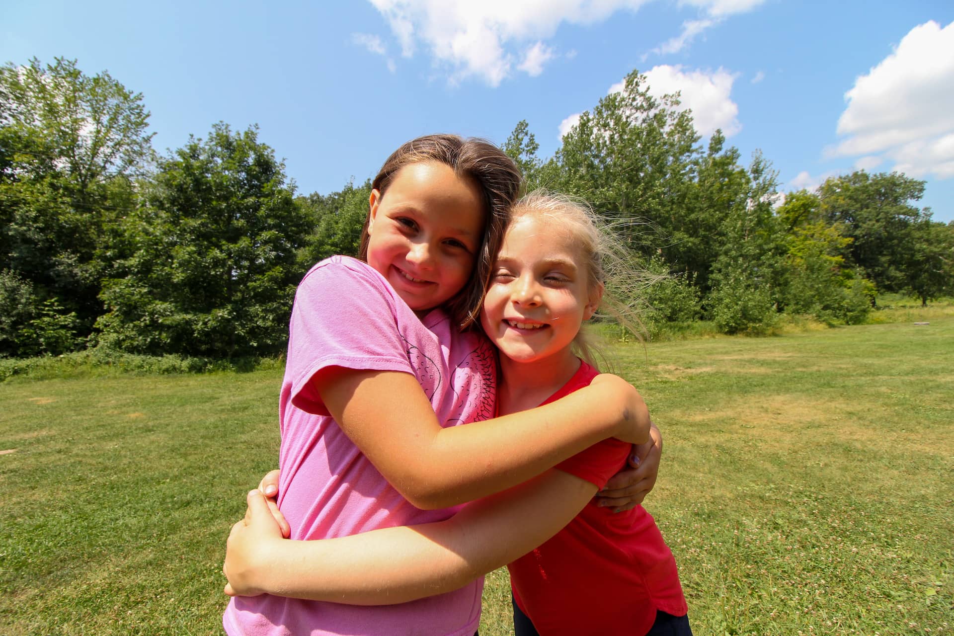 Two young girls stand outdoors hugging and smiling with trees and grass in the background under a blue sky.