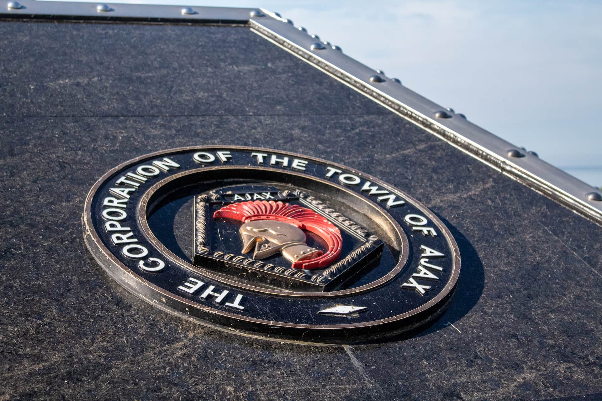 The seal of the Town of Ajax, featuring a helmet emblem, is mounted on a dark, angled surface.