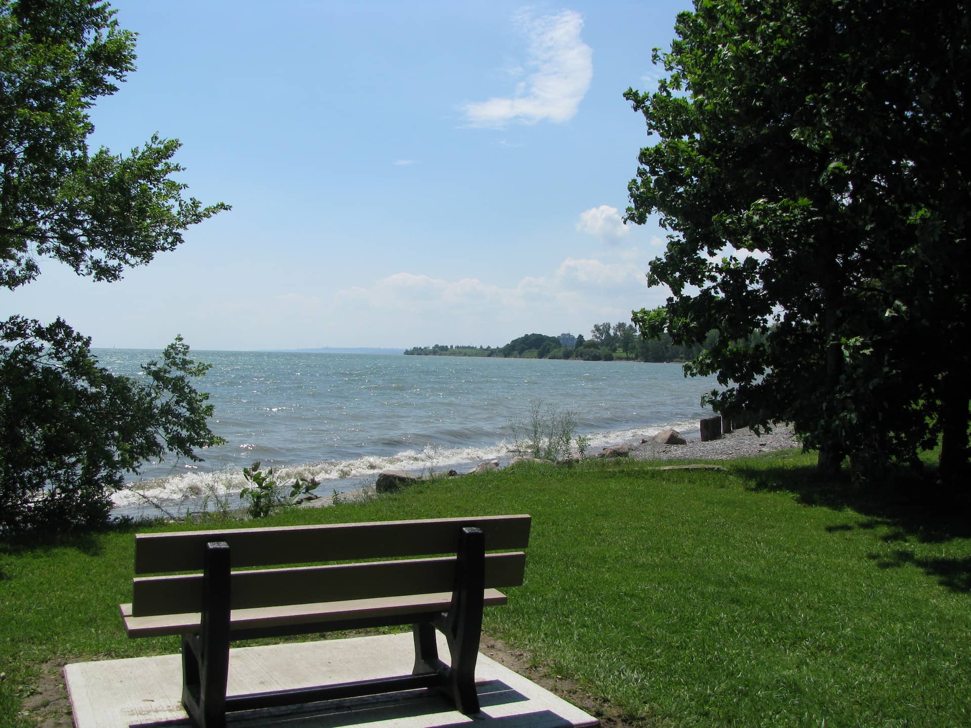 Empty bench facing a lake with gentle waves, surrounded by green grass and trees under a partly cloudy sky.