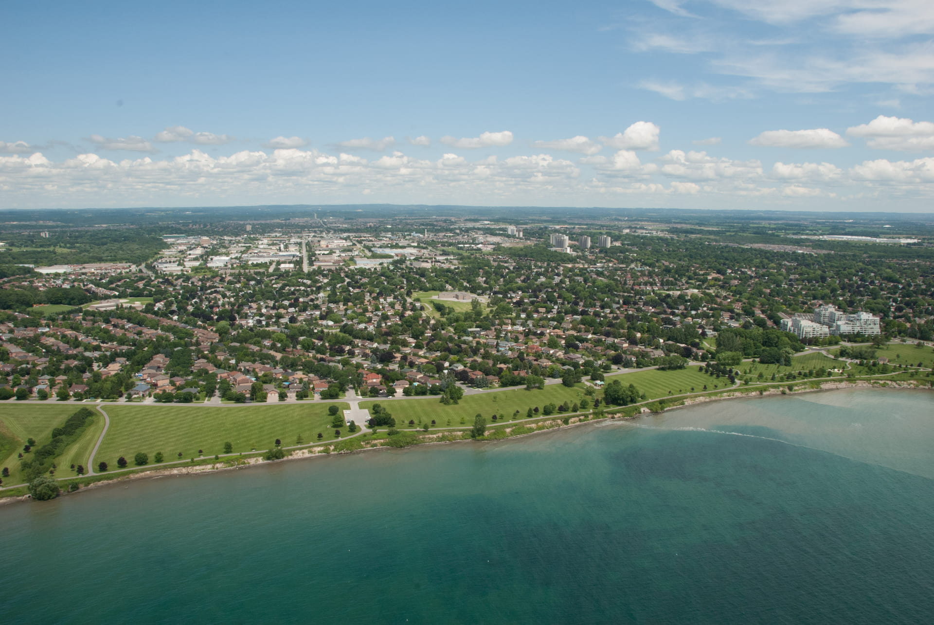 Aerial view of a suburban neighbourhood with green spaces beside a body of water under a partly cloudy sky.