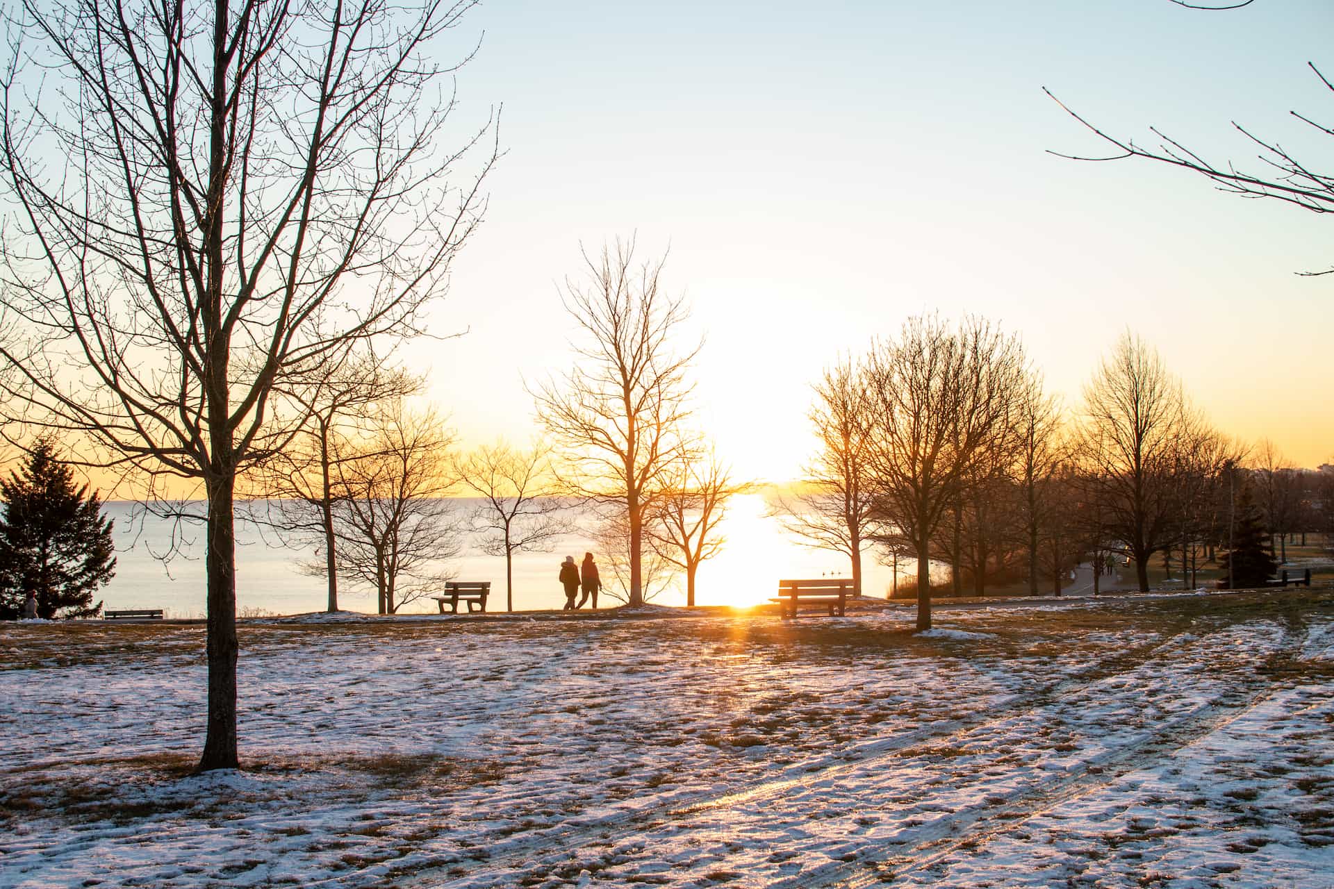 Two people walk near leafless trees in a snowy park at sunrise, with benches and a lake in the background.