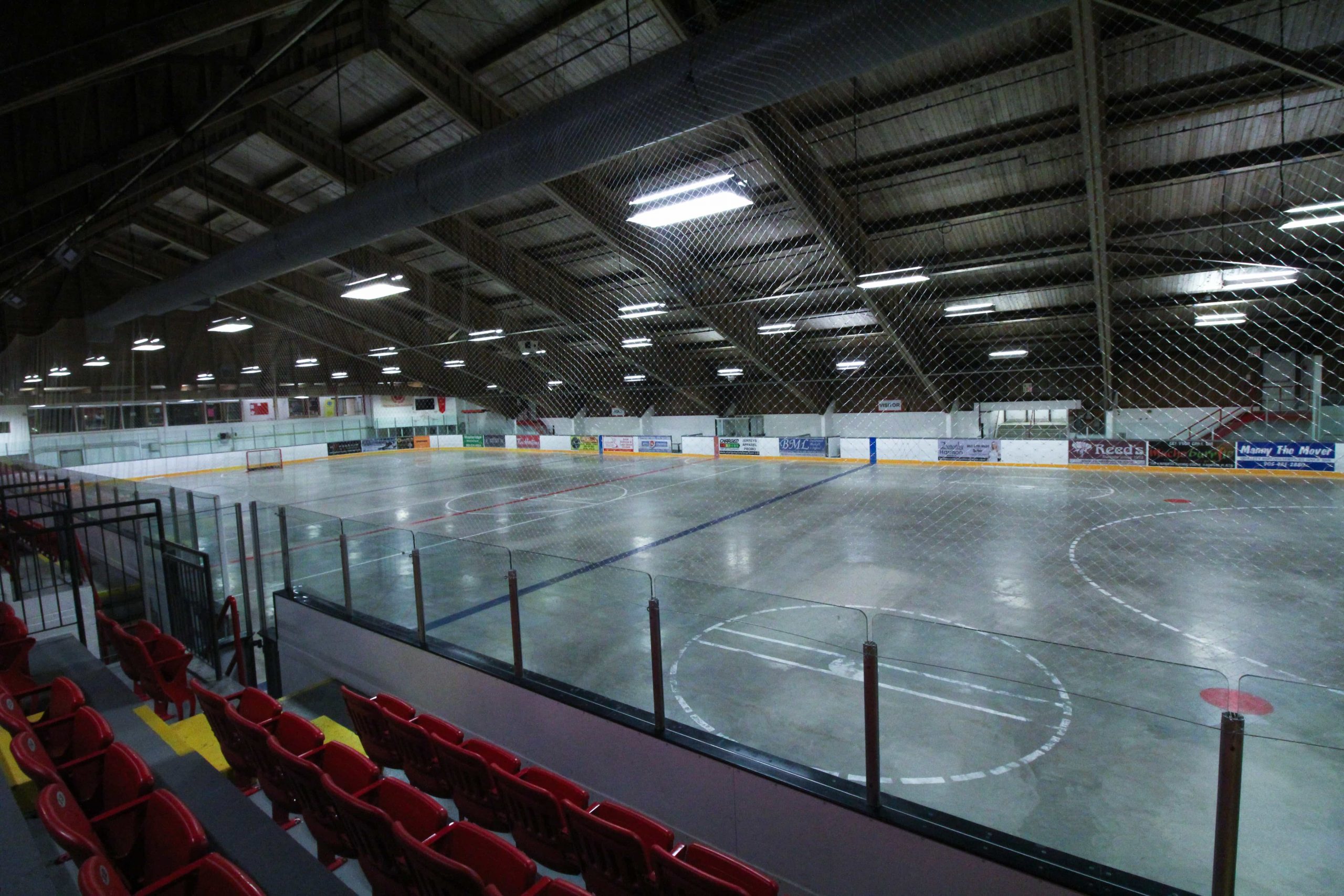 Empty indoor sports arena with red seating, surrounded by glass panels and netting, overlooks a marked concrete floor.