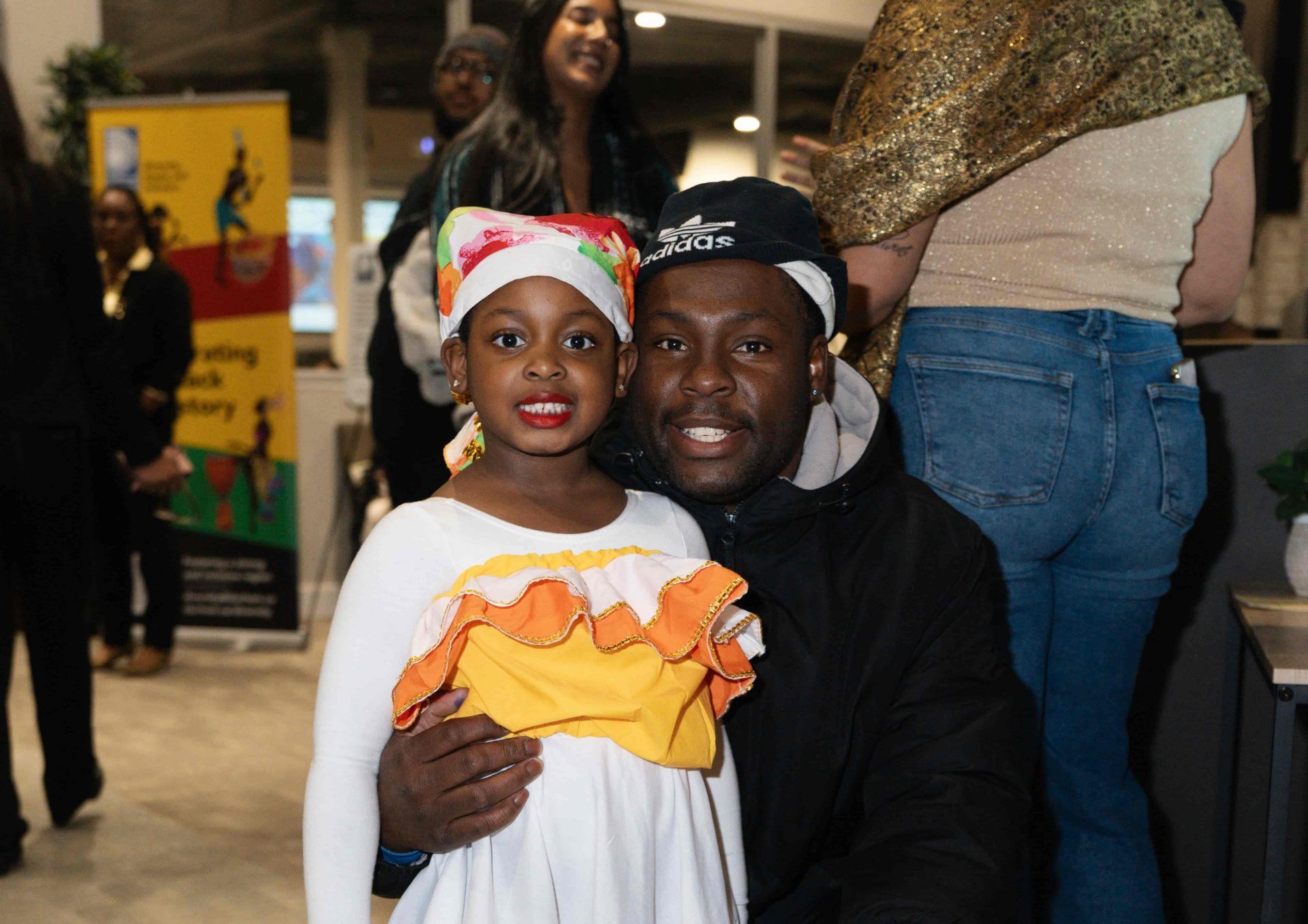 A young girl in a white dress and colourful headscarf stands with a man kneeling beside her at an indoor event.