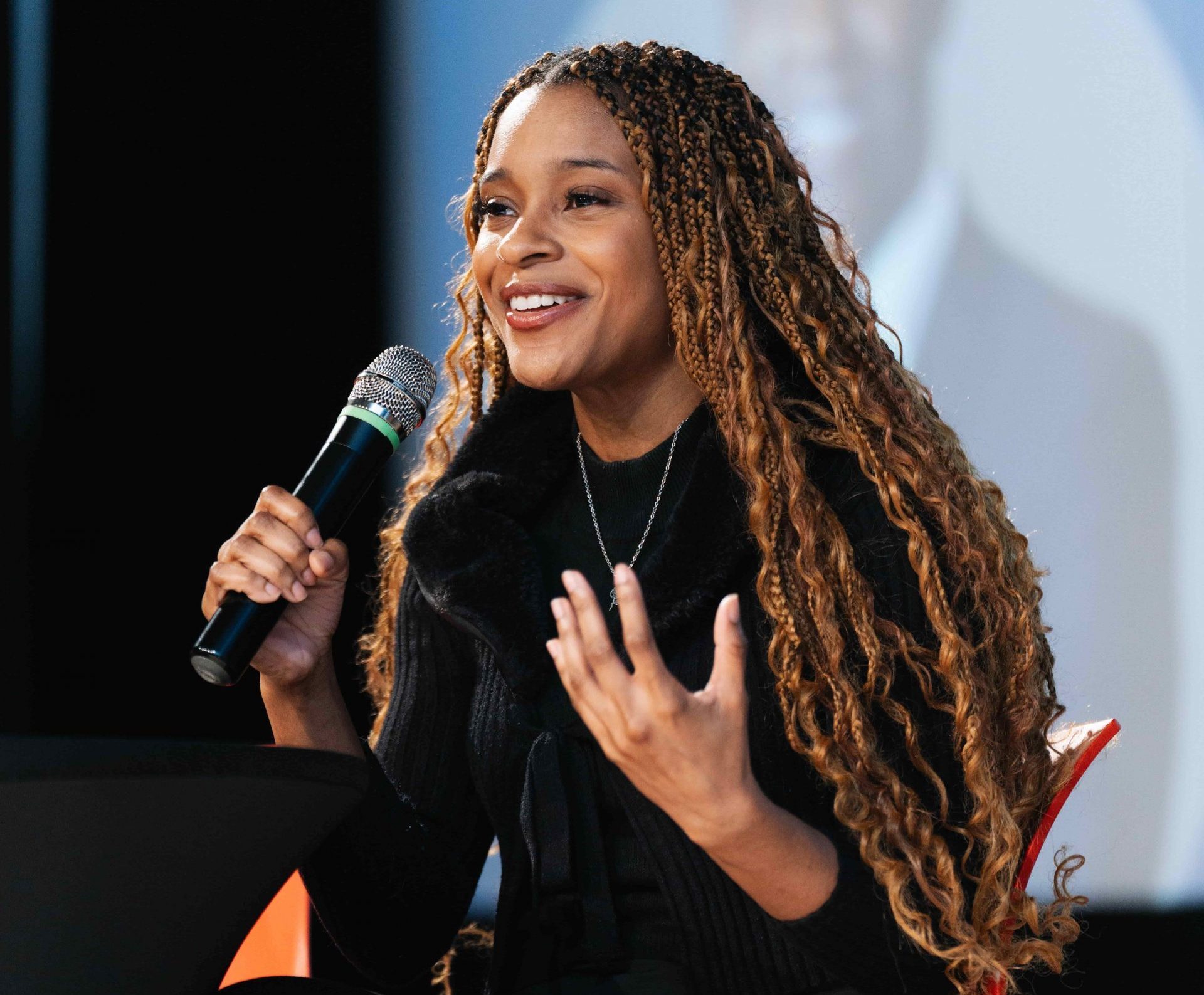Woman with long plaits sits on an orange chair, speaking into a microphone during an event or panel discussion.