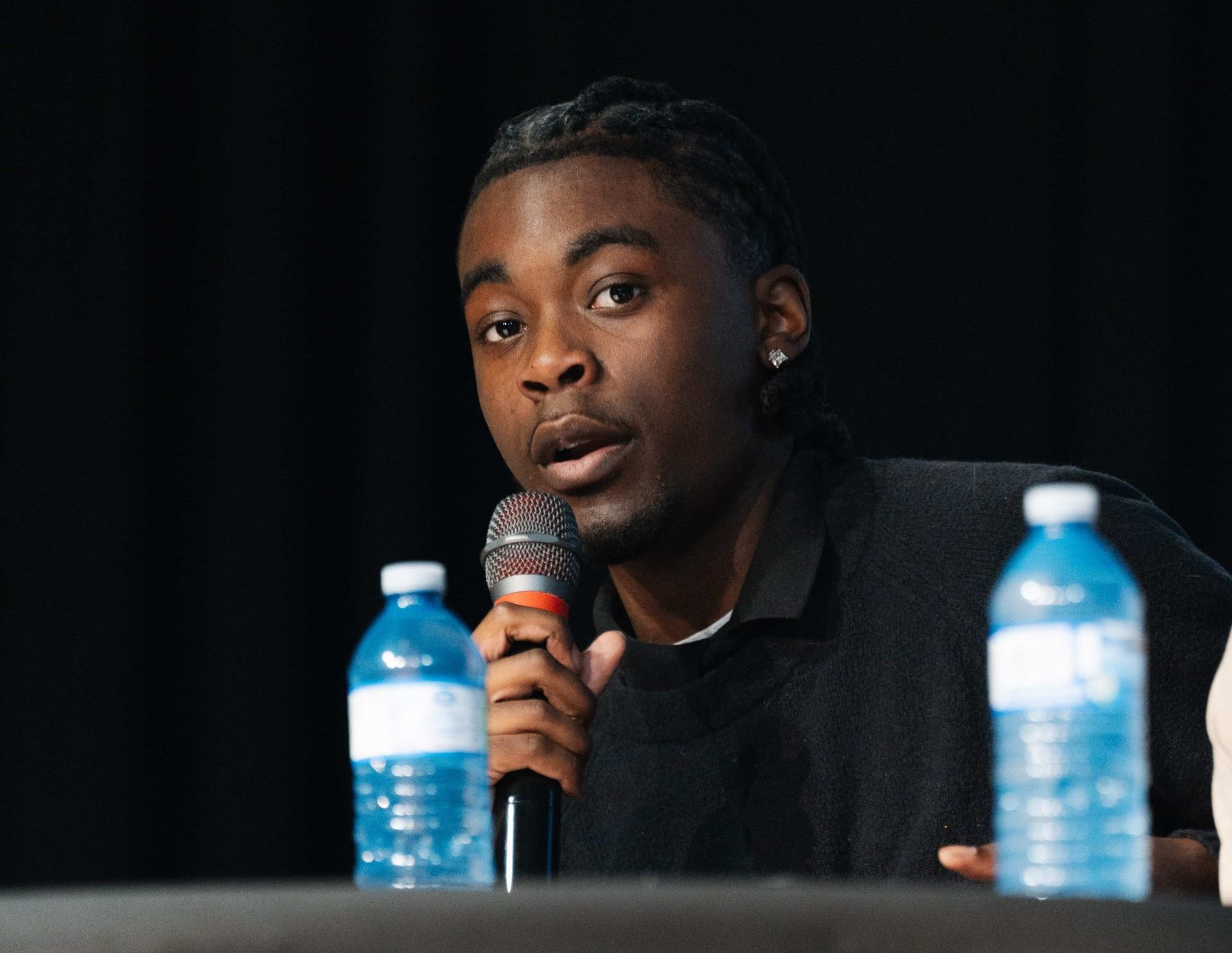 A person speaks into a microphone whilst seated at a table with two bottles of water in front of them.