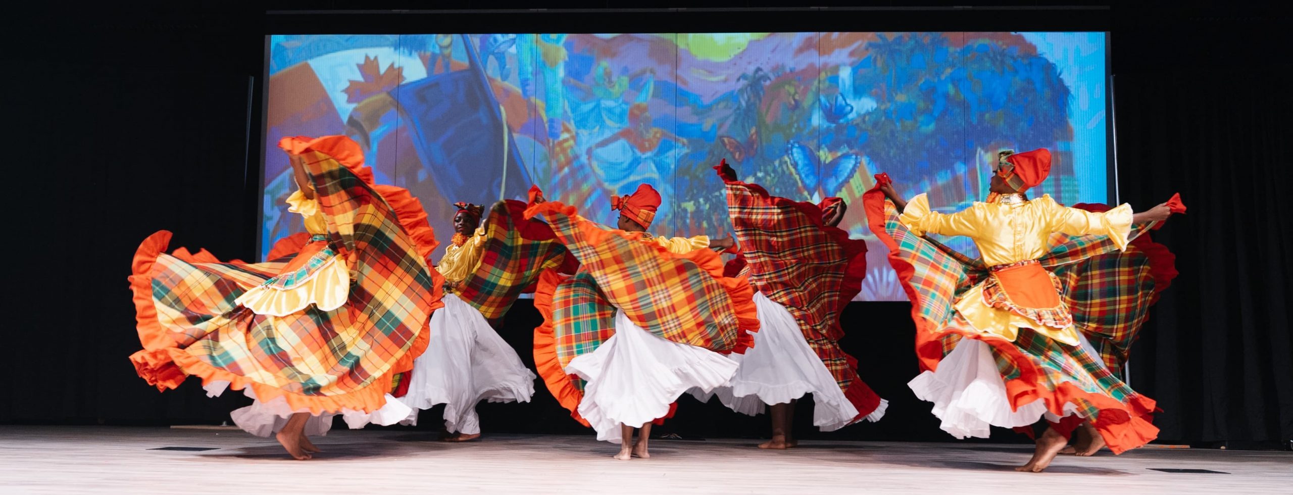 Dancers in colourful plaid costumes perform on stage with vibrant artwork projected in the background.