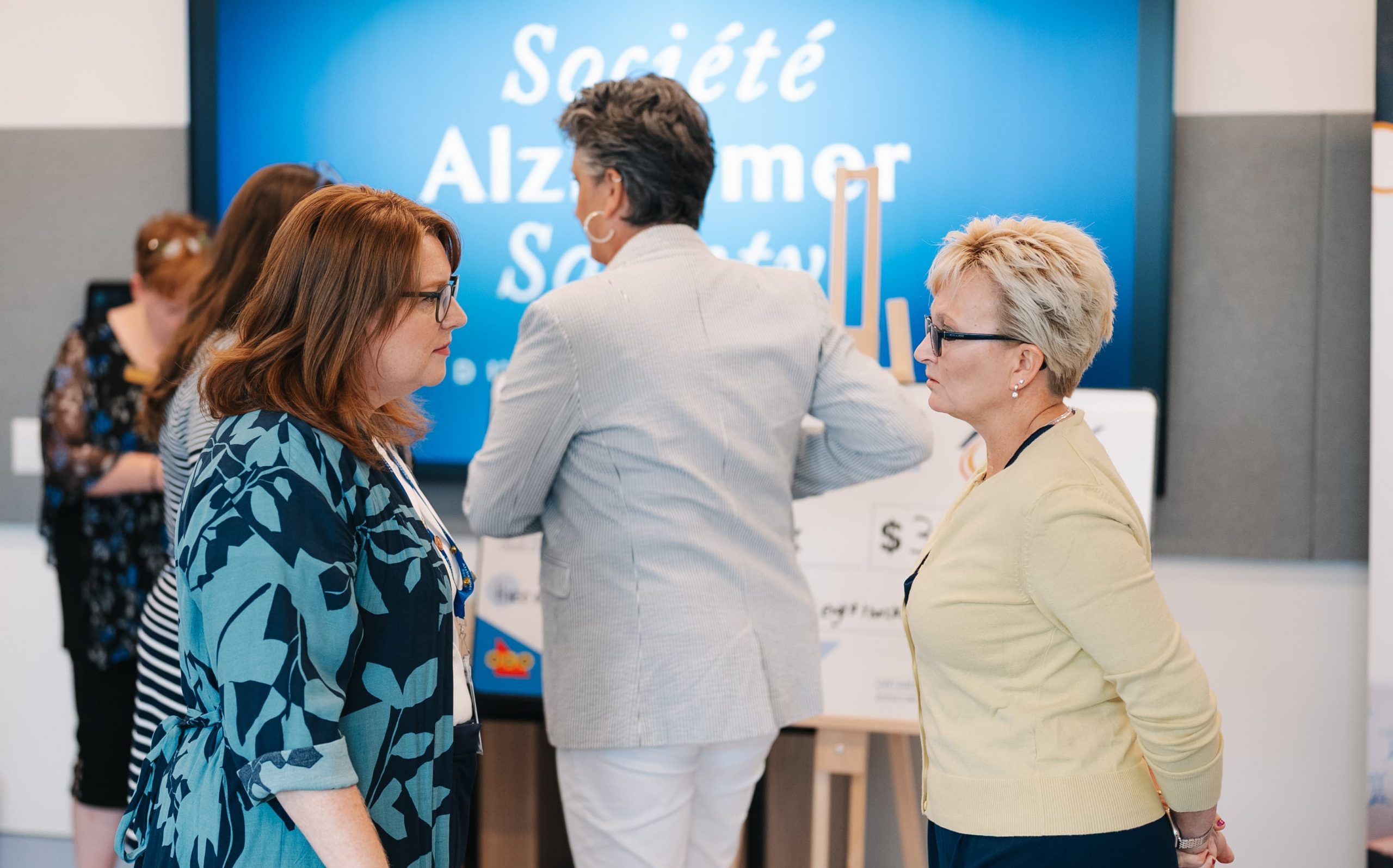 Two women are talking in a room with other people, with an Alzheimer’s Society sign in the background.