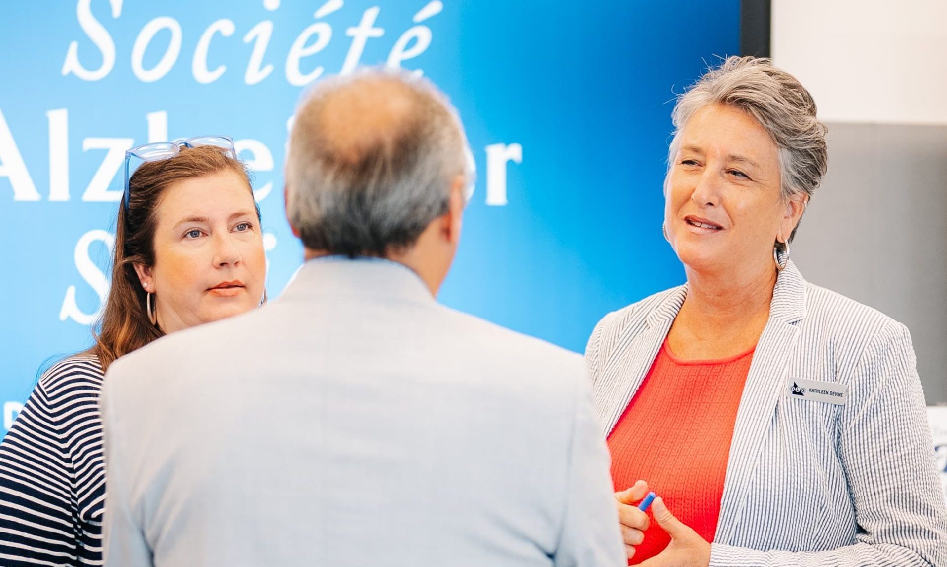 Three people are having a discussion in front of a screen displaying the text "Société Alzheimer.