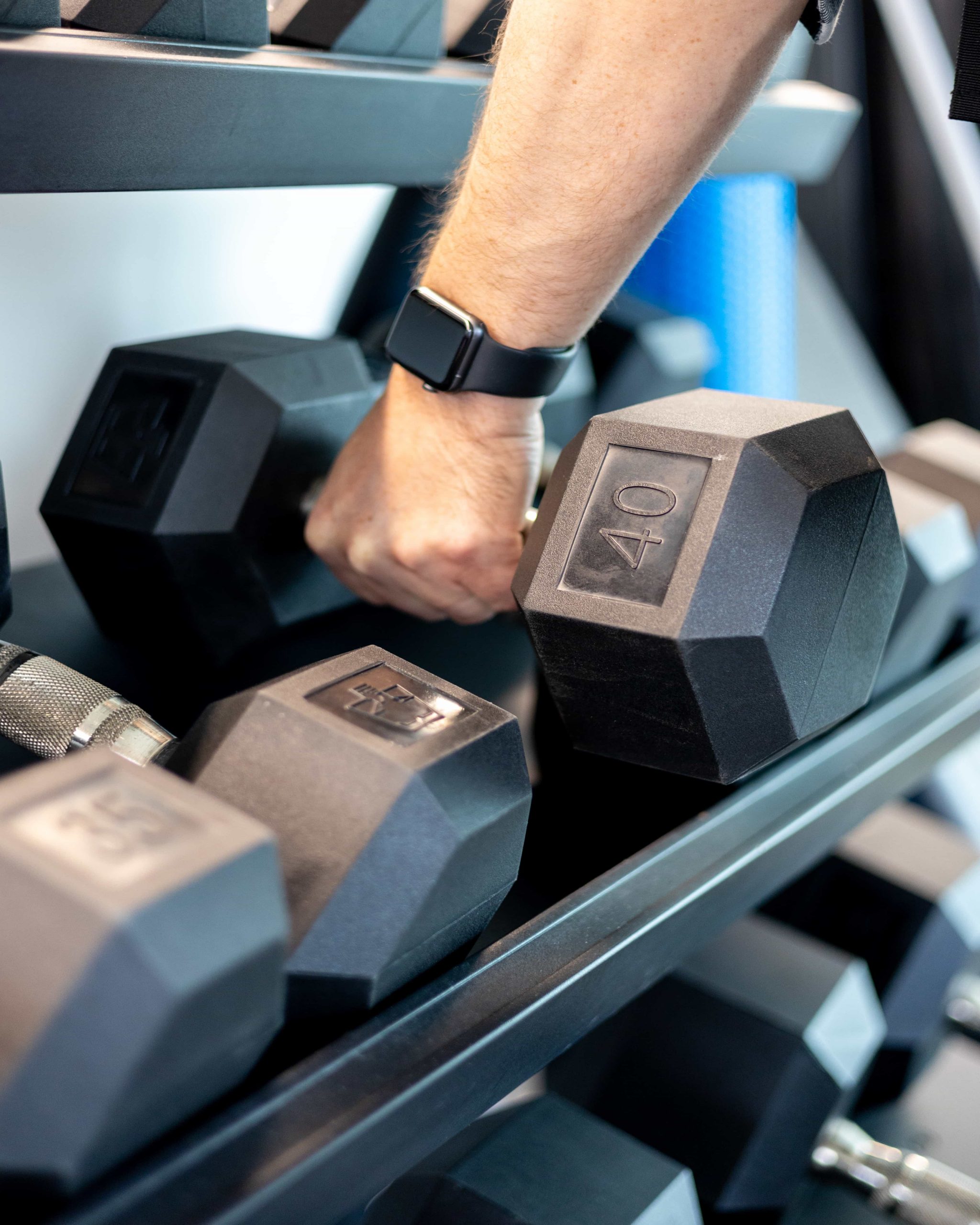 A person wearing a smartwatch lifts a 40-pound dumbbell from a rack of hexagonal weights at the gym.