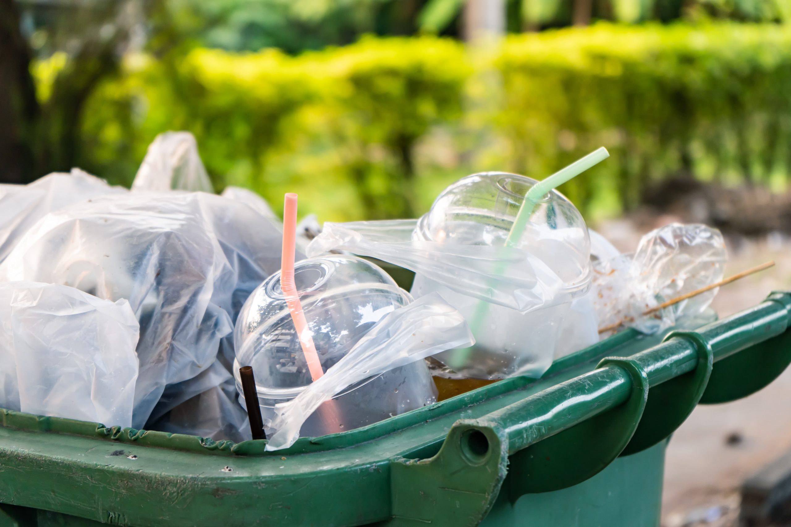 Plastic cups with straws and bags overflowing from a green rubbish bin outdoors, with greenery in the background.