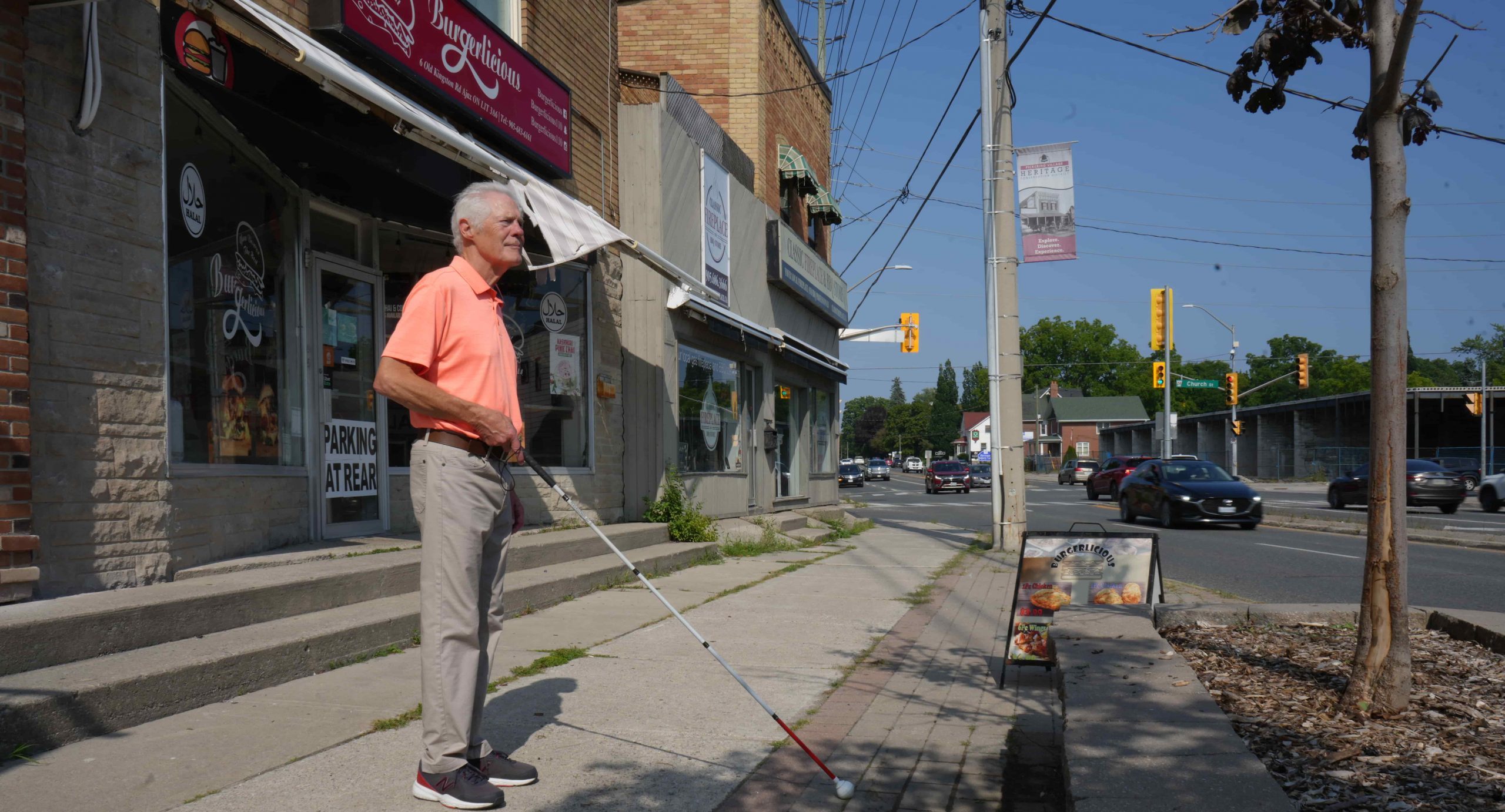 An older man with a white cane stands on a city pavement near shops, appearing to navigate the street.
