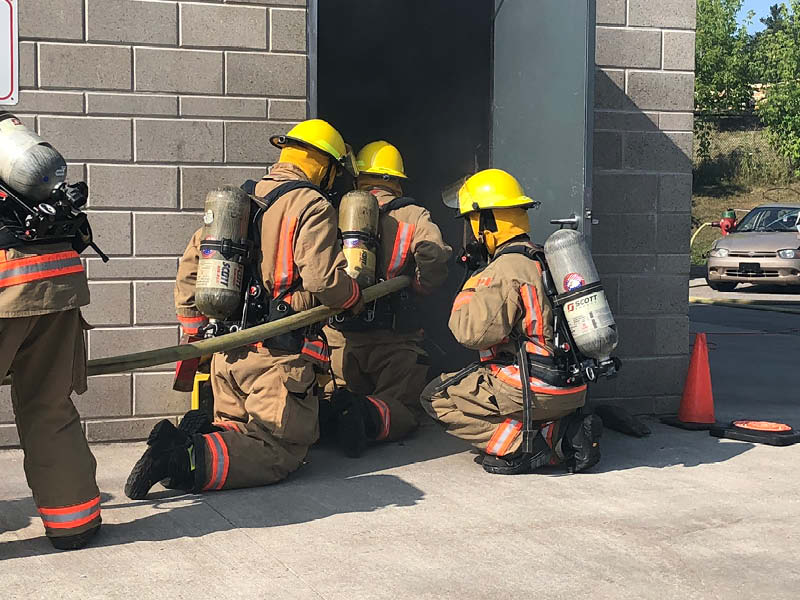 Three firefighters in gear kneel at an open door with a hose, preparing to enter a smoky building.