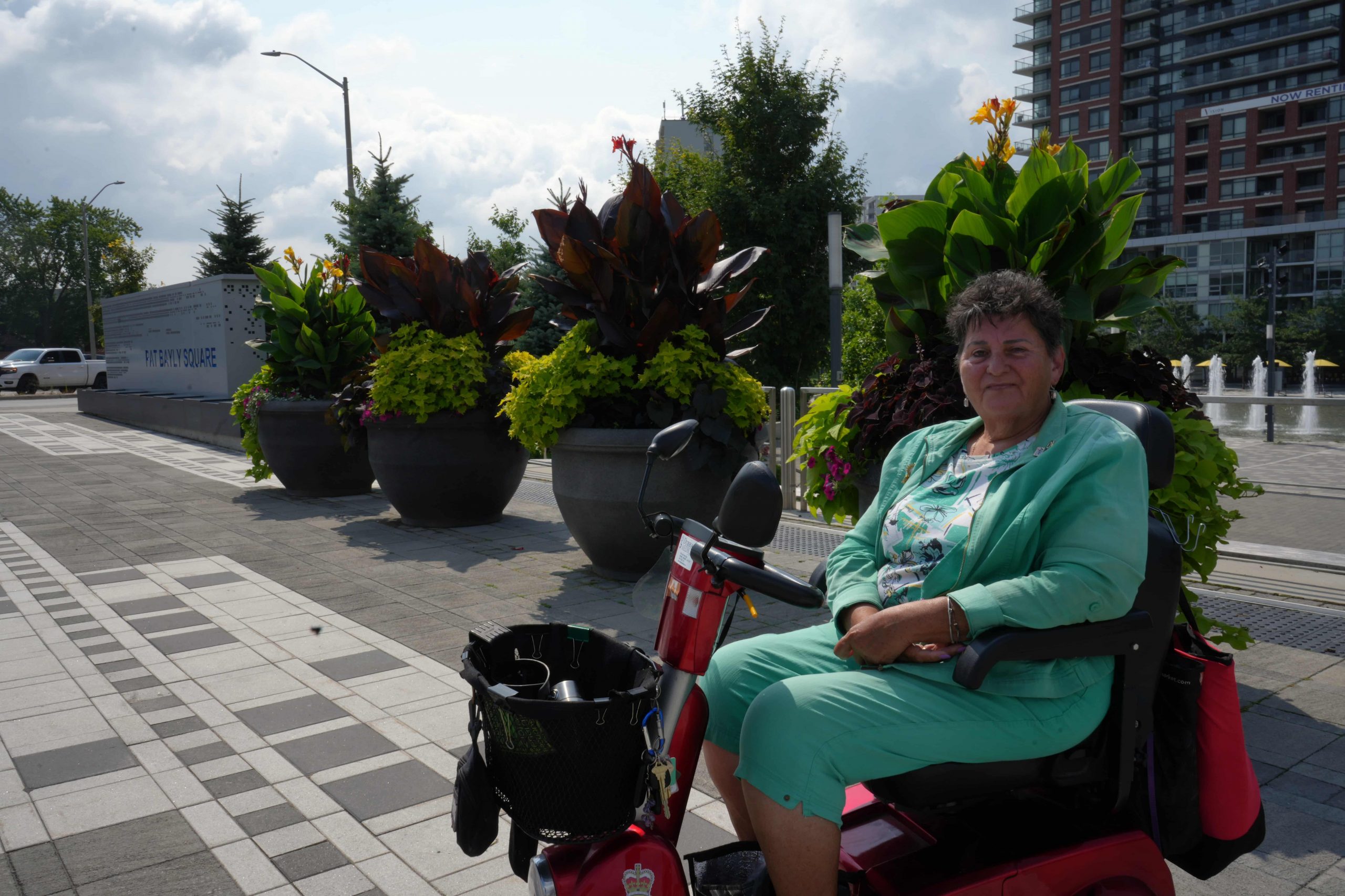 A woman in a green outfit sits on a mobility scooter near large plant pots in an urban square.