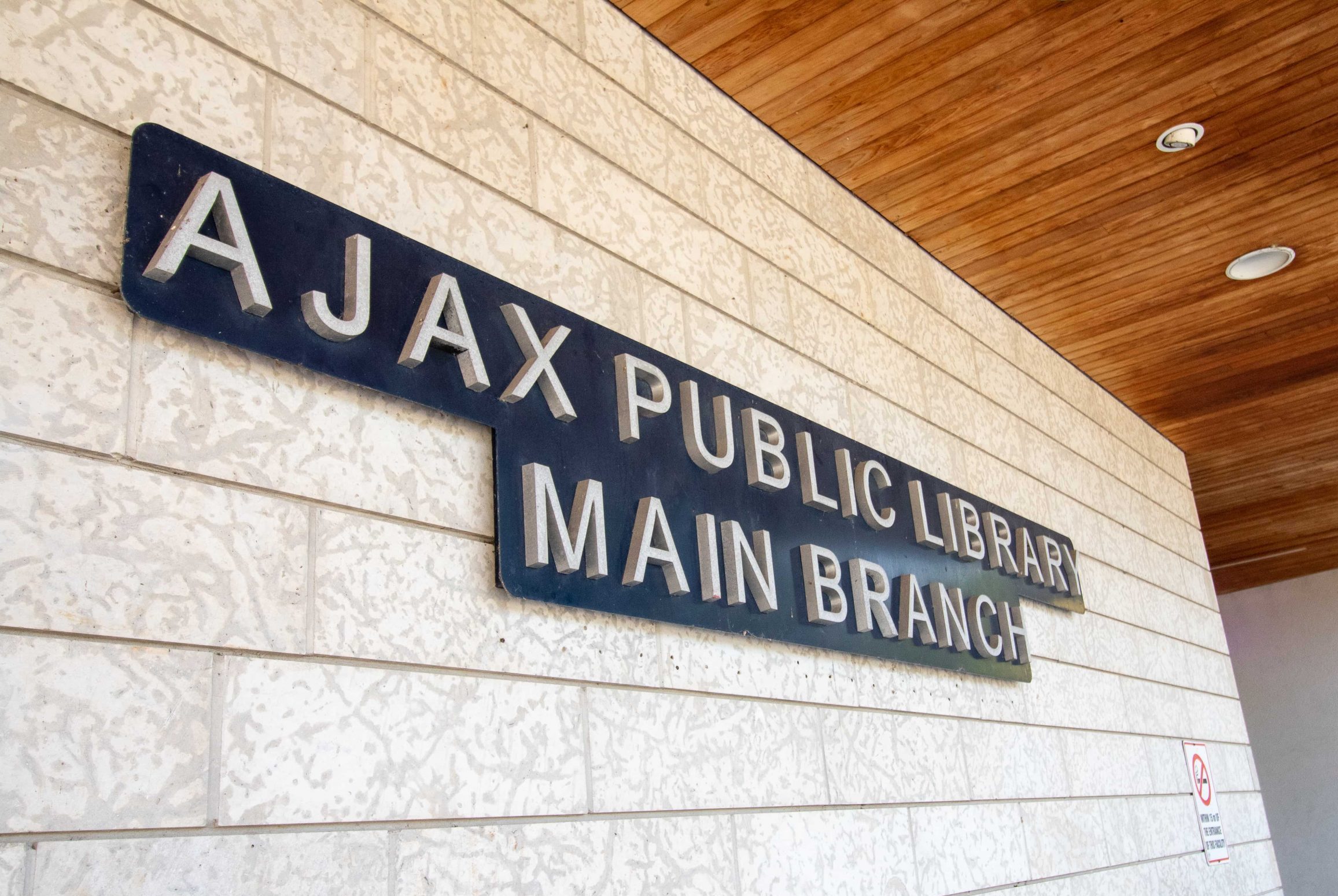 Sign for Ajax Public Library Main Branch mounted on a light-coloured brick wall under a wooden overhang.