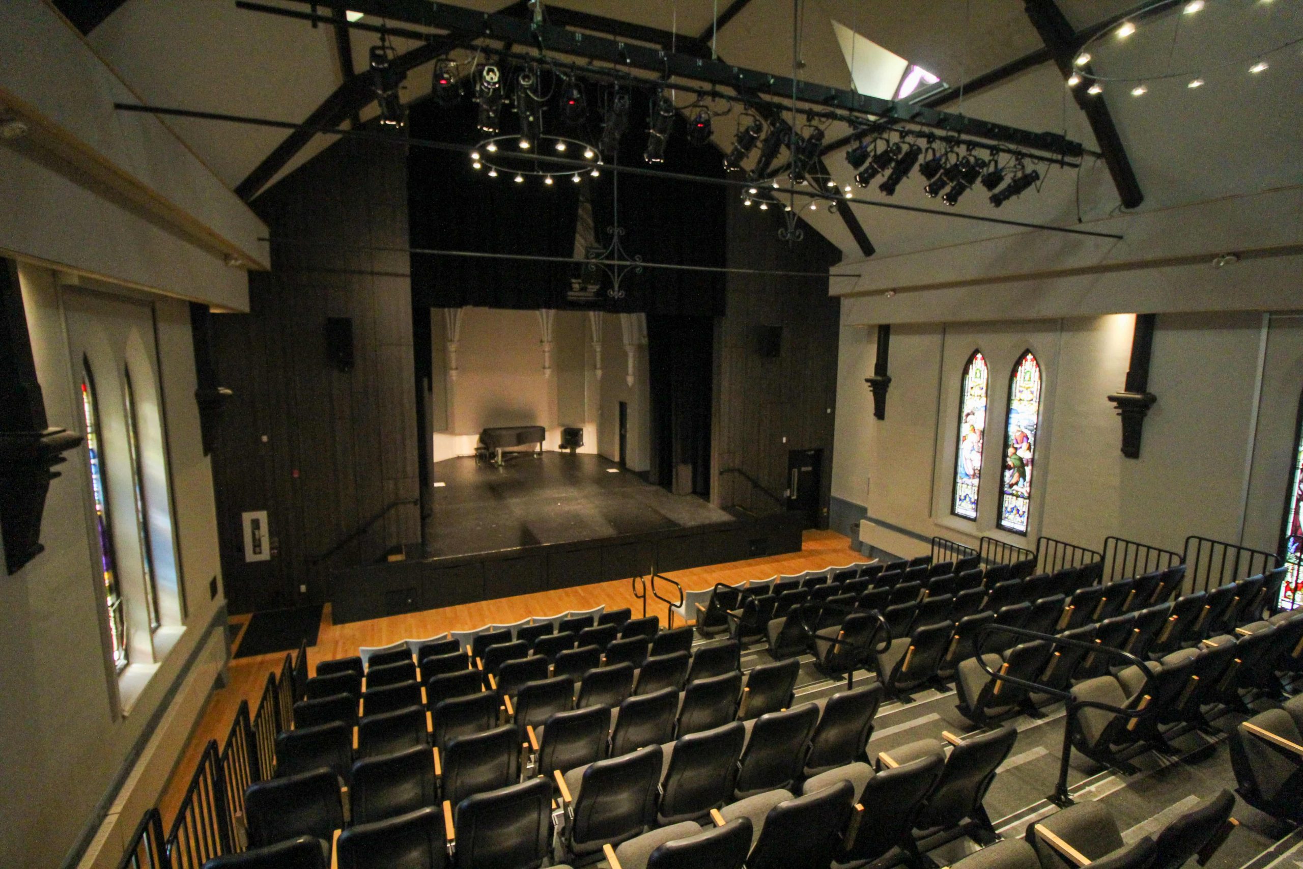 An empty theatre with rows of seats facing a stage, overhead lighting, and stained glass windows on the side walls.