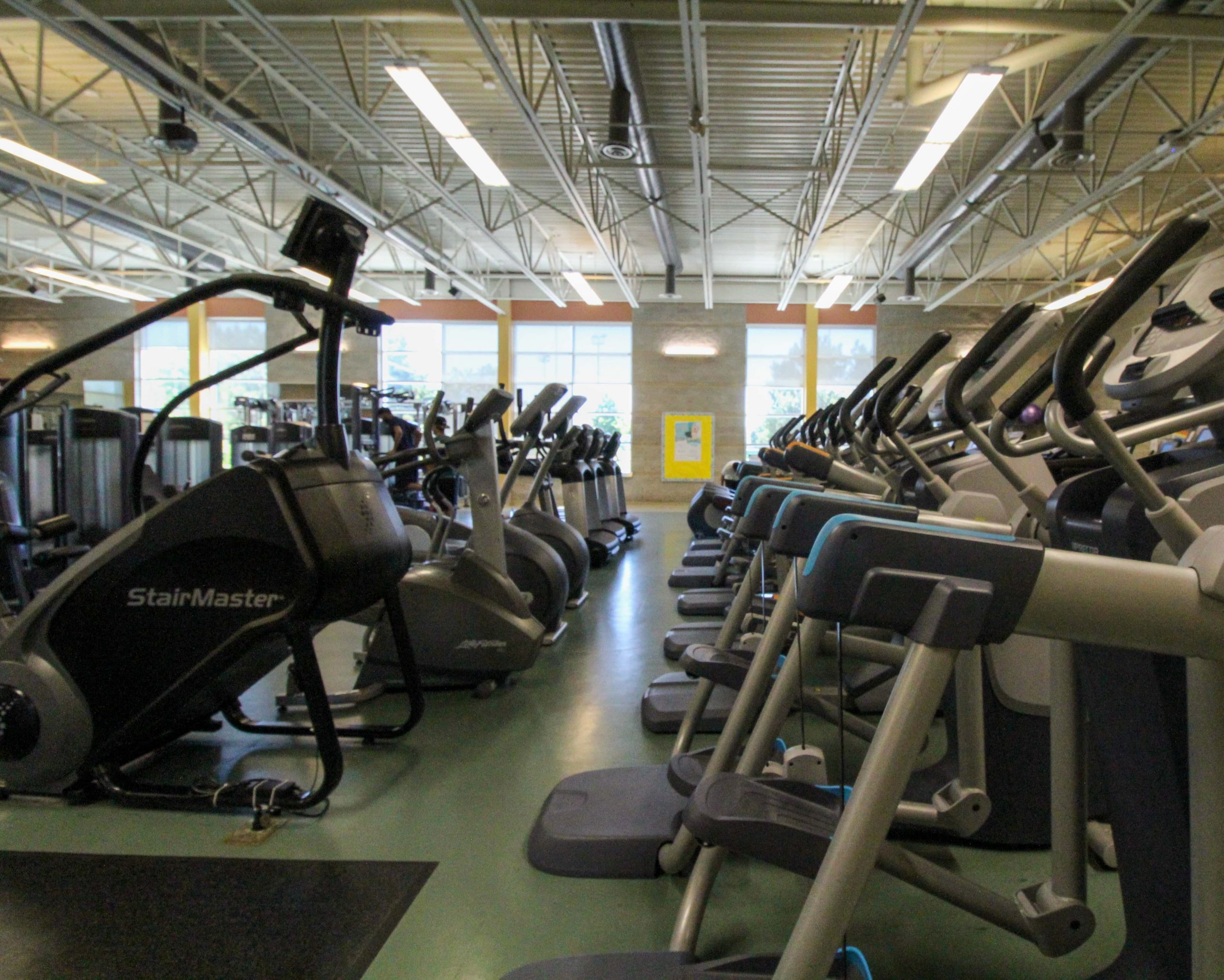 Rows of exercise machines, including stairmasters and treadmills, are lined up in a well-lit gym with large windows.