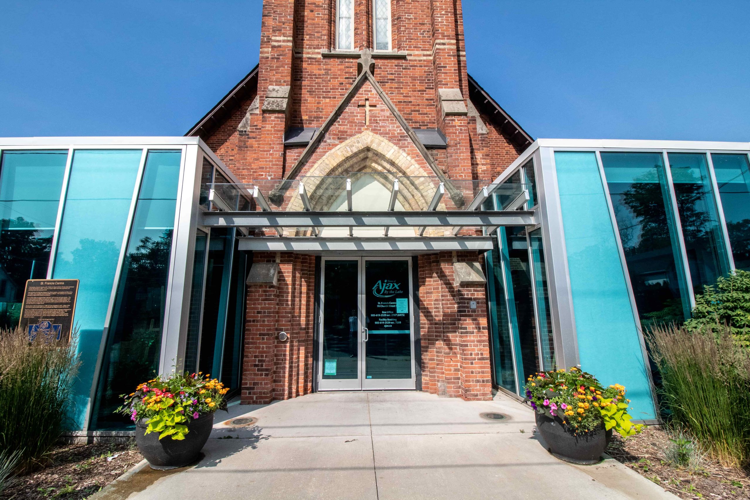 Glass entrance to a brick building with potted plants on either side and a sign reading "Ajax Public Library".