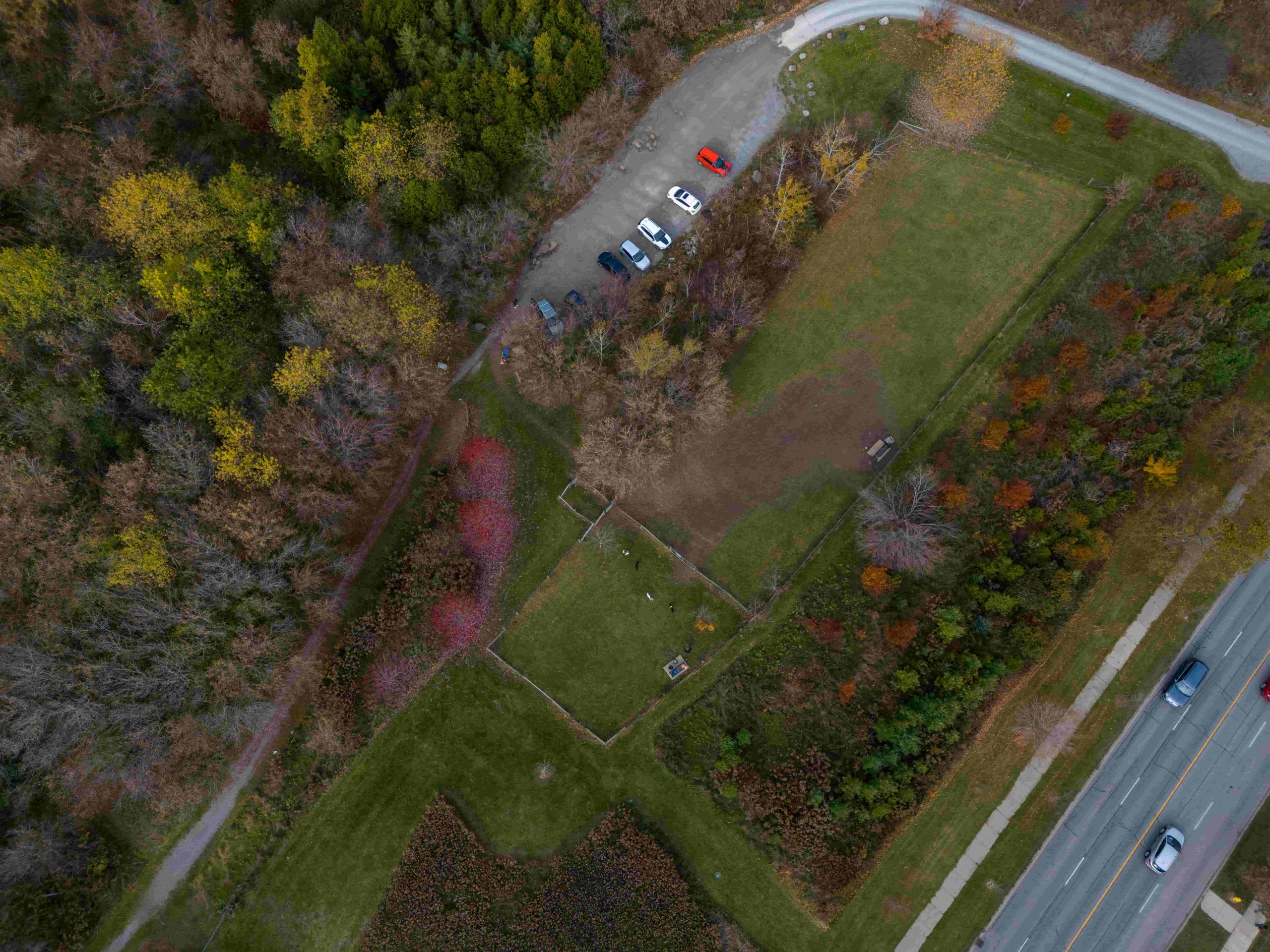 Aerial view of a car park with cars, grassy fields, trees, and a road with vehicles on the right side.
