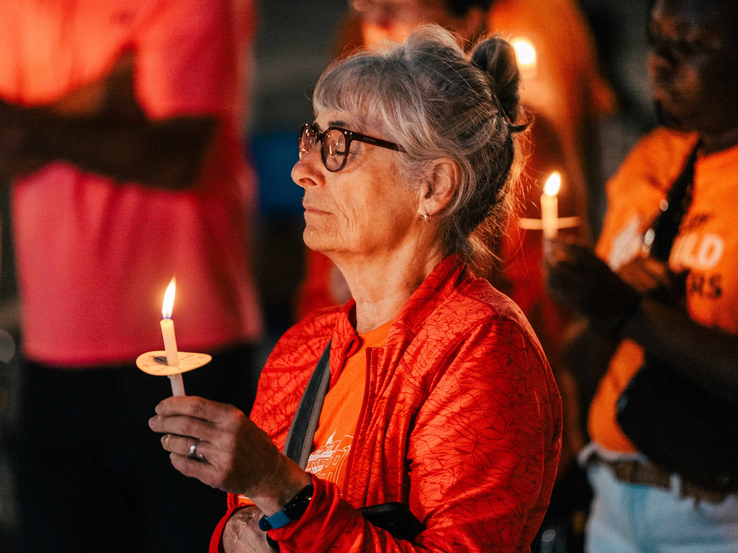 An older woman holding a lit candle stands among others at a vigil or memorial event.