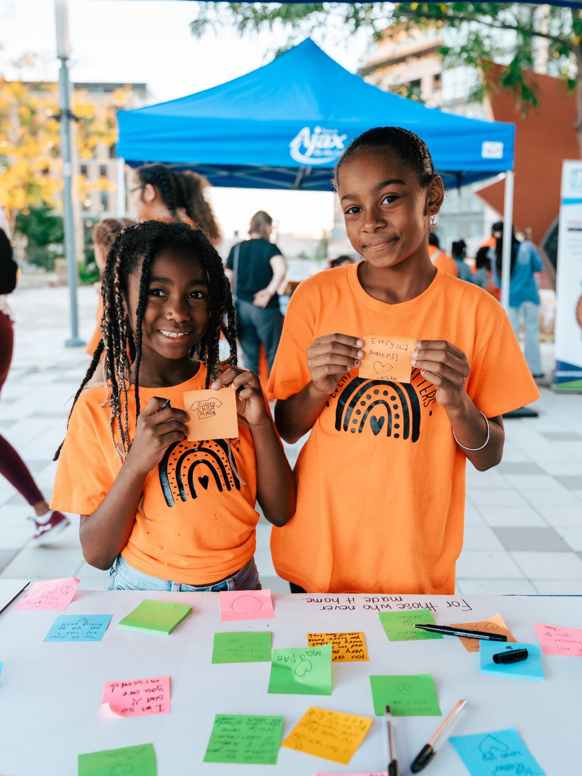 Two children in orange shirts hold up cards at a table covered with colourful notes at an outdoor event.