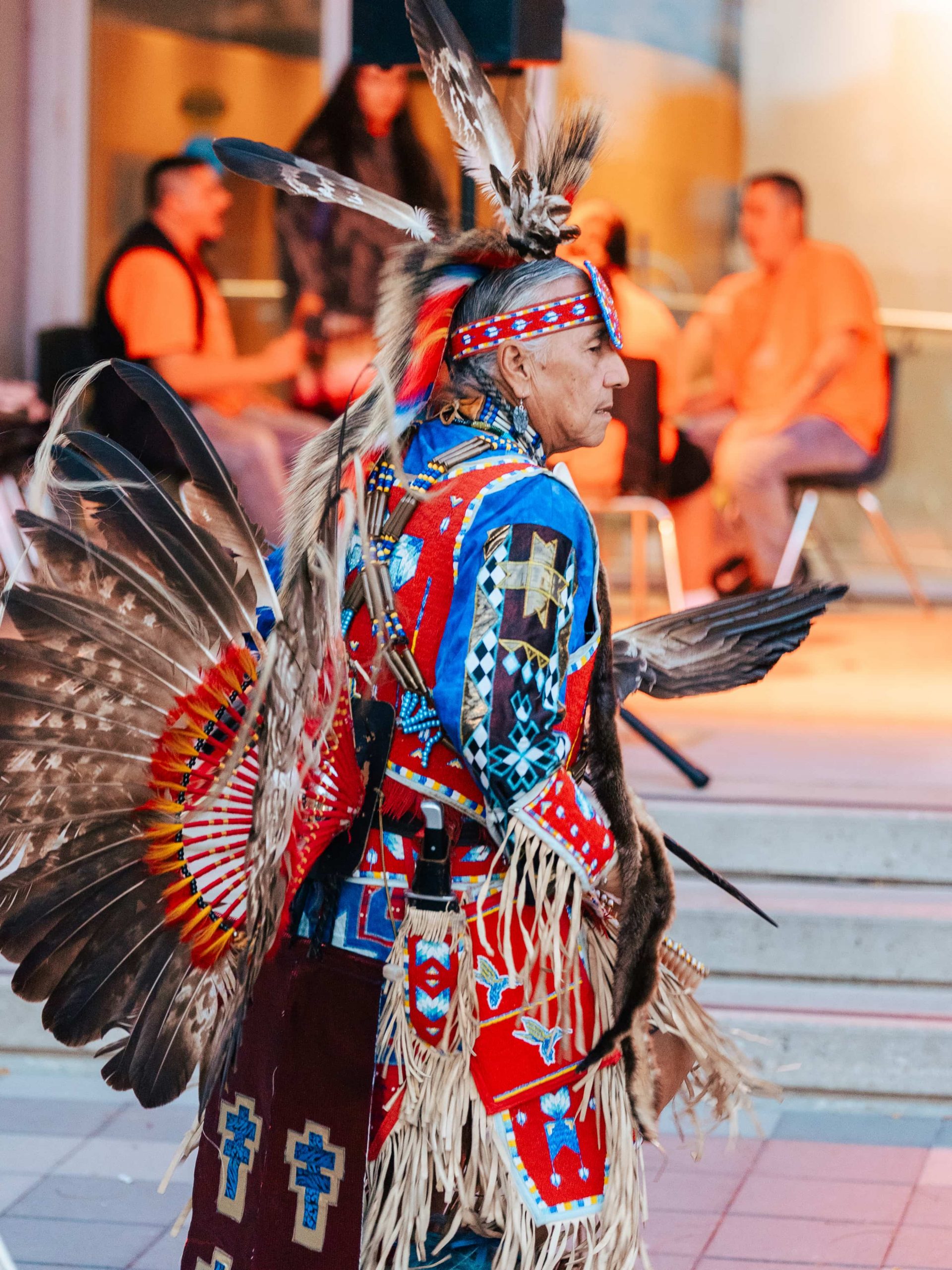 A person in traditional Native American regalia dances outdoors whilst others sit and watch in the background.