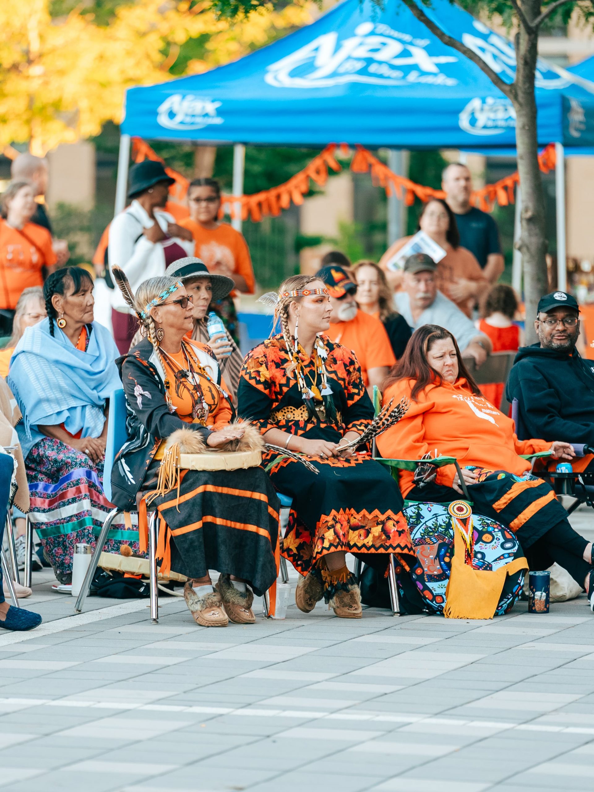 A group of people in traditional and orange clothing are seated outdoors at an event with an Ajax marquee in the background.