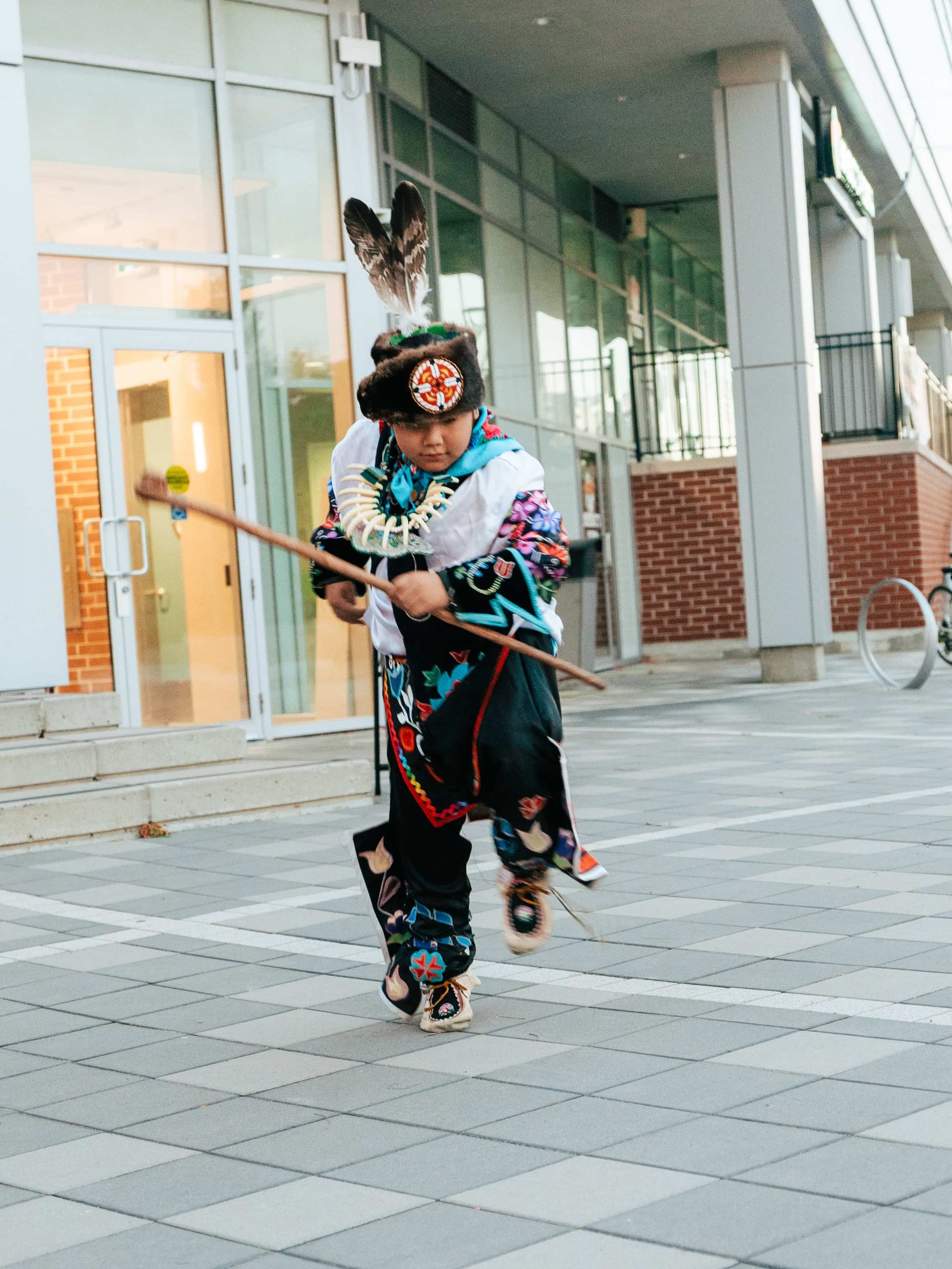 A person in traditional Native American attire performs a dance outdoors on a tiled pavement near a modern building.