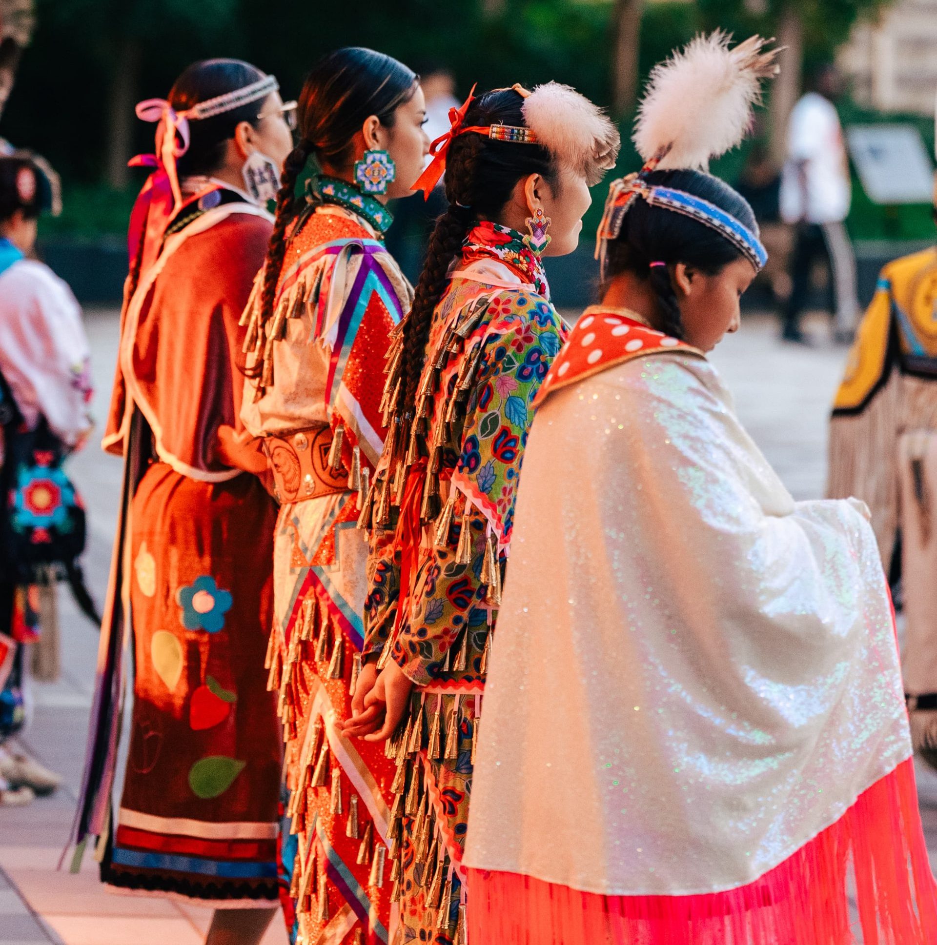 A group of people in colourful traditional regalia stand outdoors in a line, facing away from the camera.