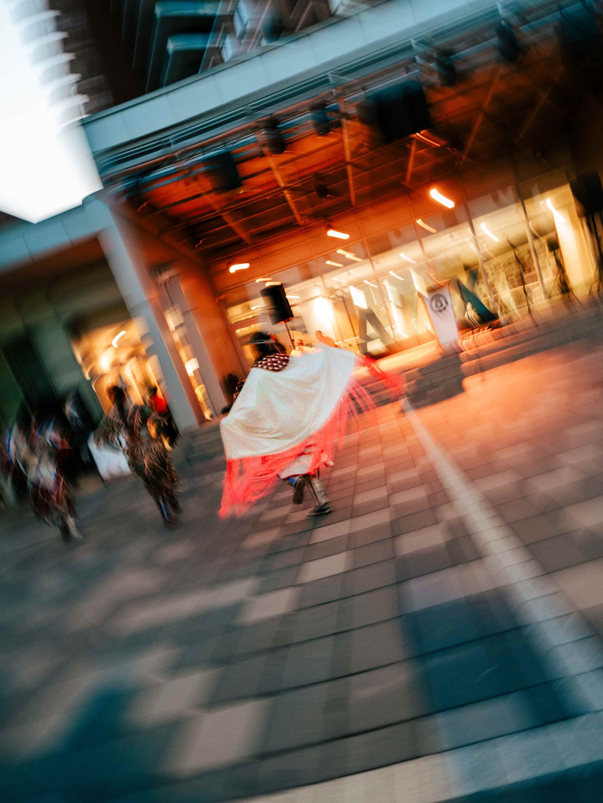 A person in a white shawl with pink fringe dances outside a modern building, with motion blur in the image.